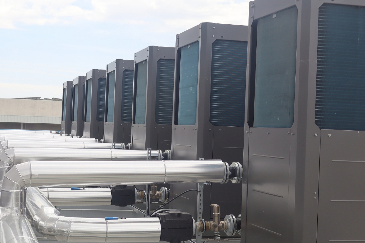 Row of industrial heat pumps connected by metal piping on the rooftop of a facility.