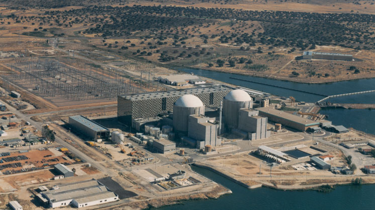 Vista aérea de una central nuclear junto a un río con edificios industriales y subestación eléctrica.