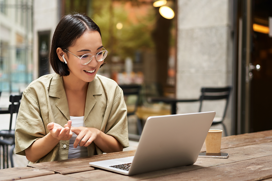 A smiling person is participating in a video call from a café, wearing wireless earphones and using a laptop. They are seated at a wooden table, with a cup of coffee and a mobile phone on the table. The person is wearing a light-colored shirt and has short hair. The background shows a modern setting with soft lighting and seating.