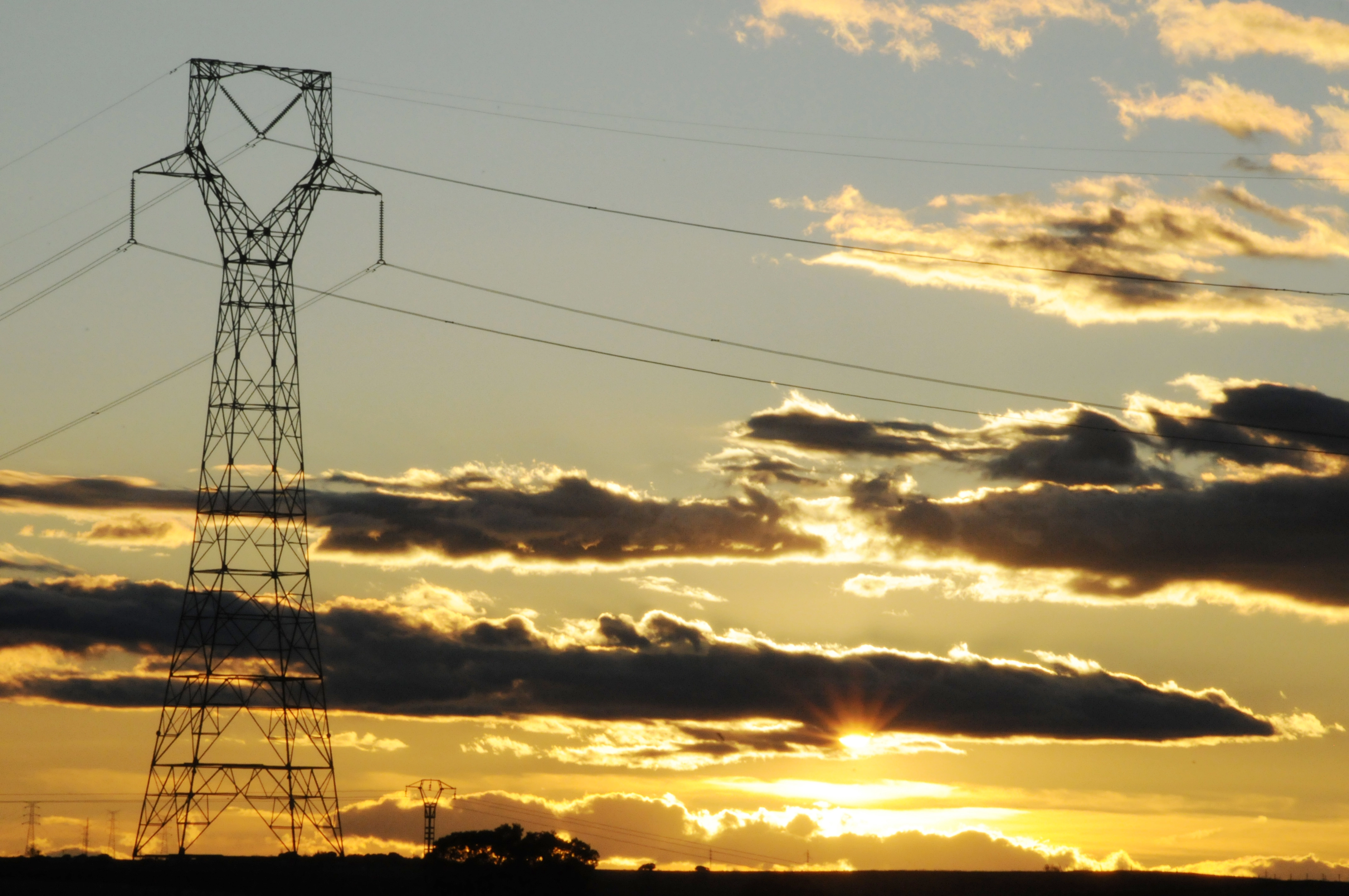 Torre de transmisión eléctrica al atardecer, con cables suspendidos y un cielo lleno de nubes iluminado por la puesta de sol.