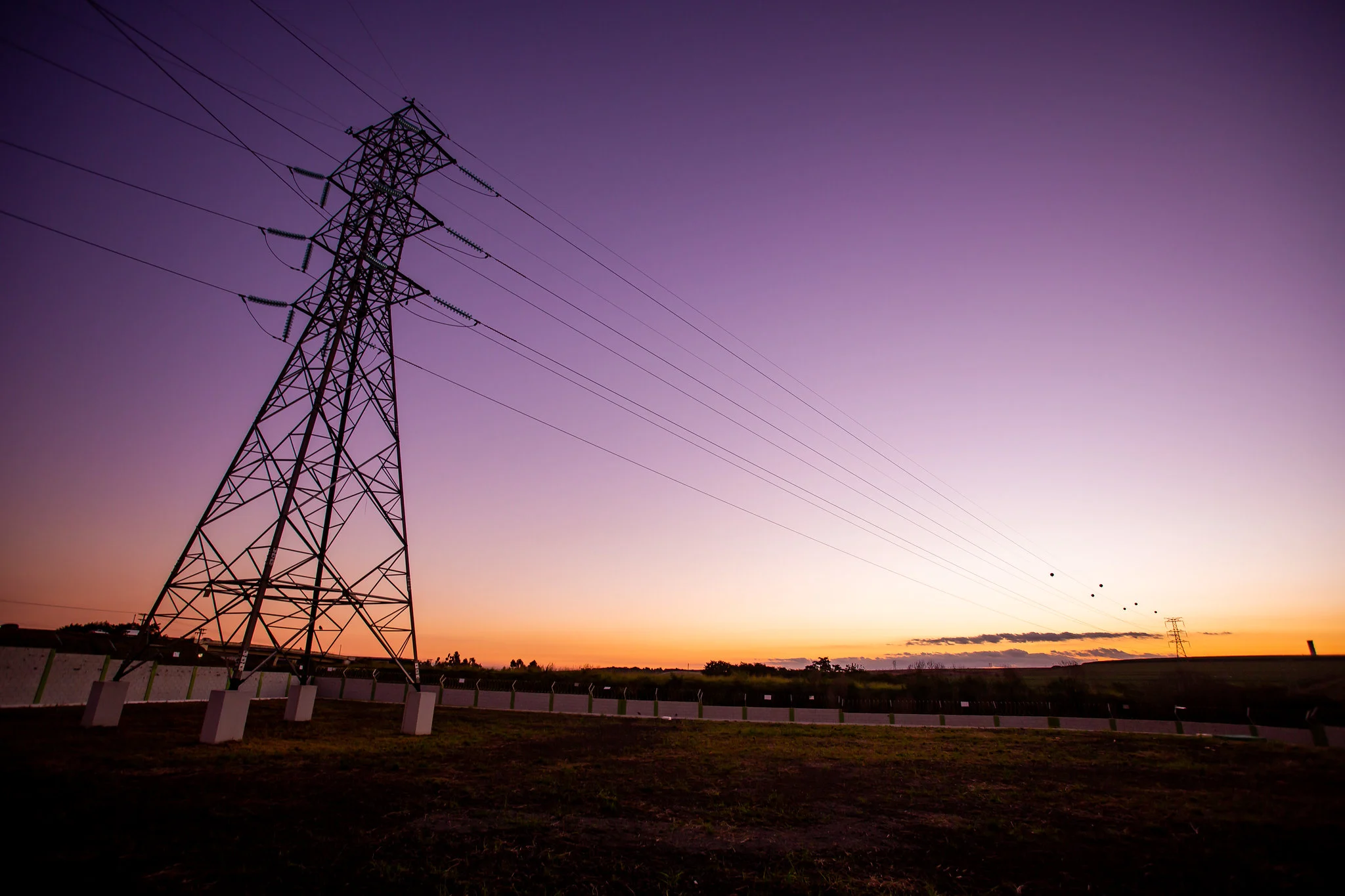 Electrical transmission tower at sunset, with cables extending toward the horizon in a purple sky.