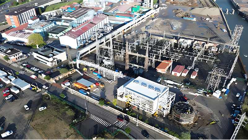 Vista aérea de una planta industrial en construcción, con infraestructuras eléctricas y vehículos estacionados alrededor.