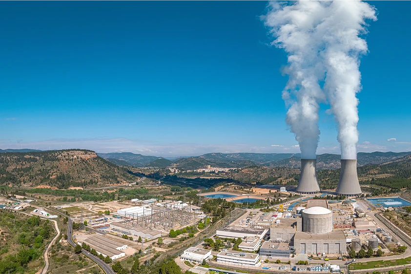 Vista aérea de una central nuclear con torres de refrigeración y vapor en un entorno montañoso.