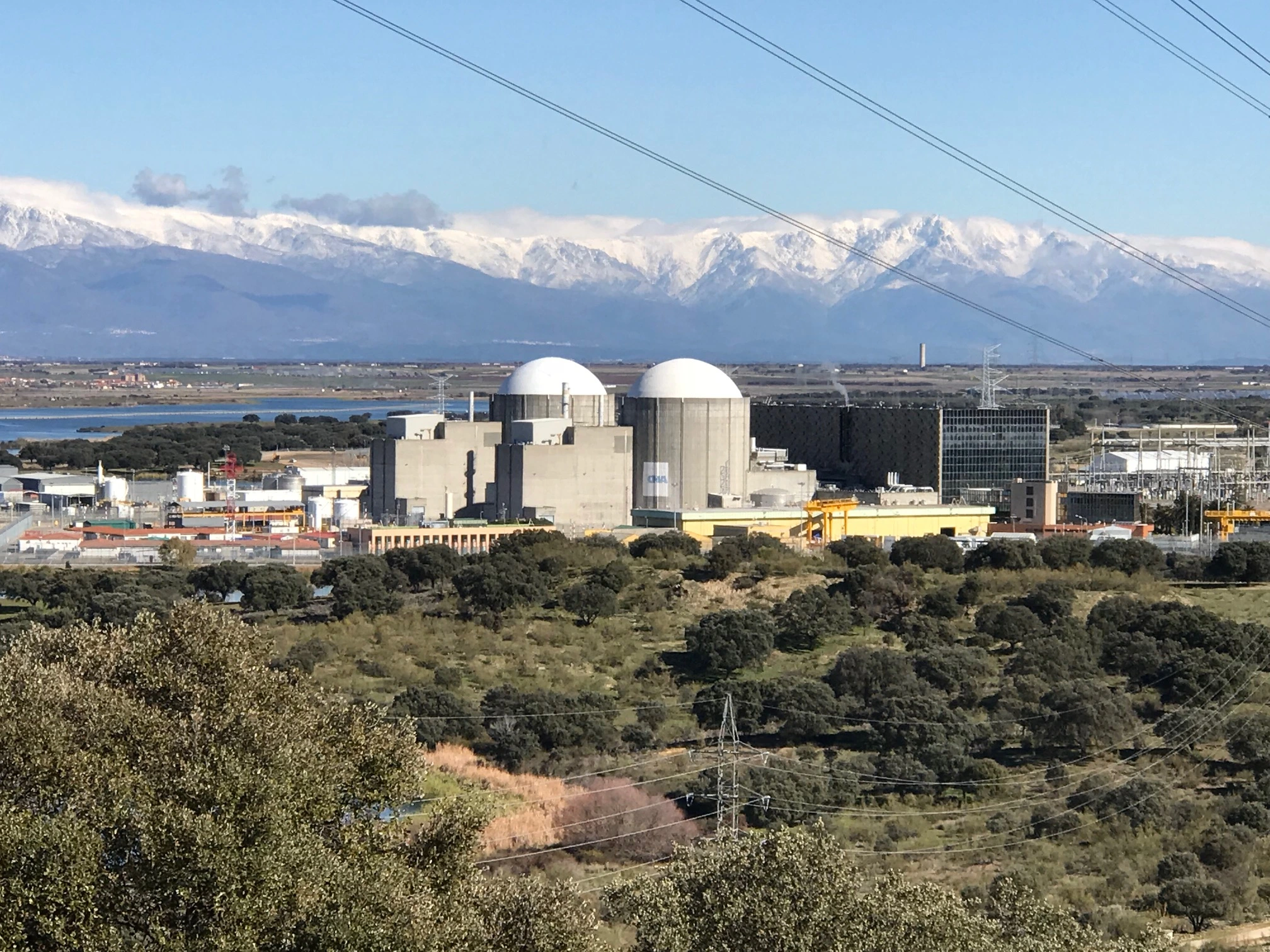 Nuclear power plant with industrial buildings and containment domes in the foreground, set against a mountainous landscape with snow-capped peaks.
