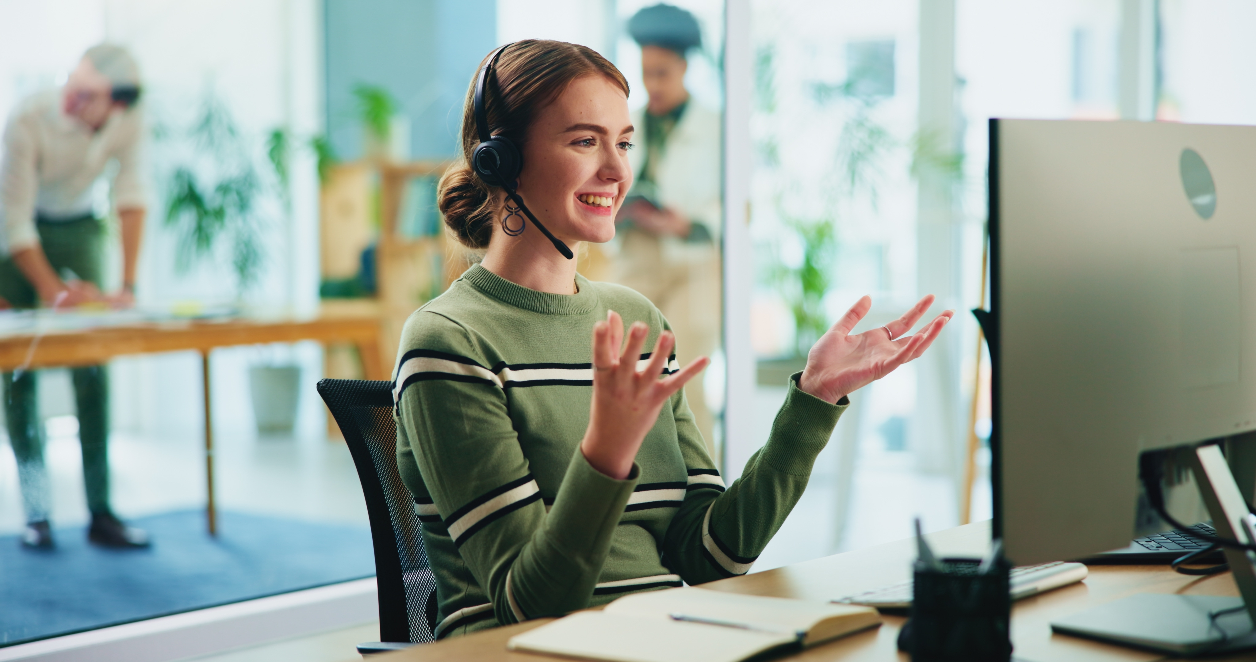 Mujer joven sonriente con auriculares y micrófono trabajando en atención al cliente desde su escritorio en oficina moderna con compañeros desenfocados al fondo.