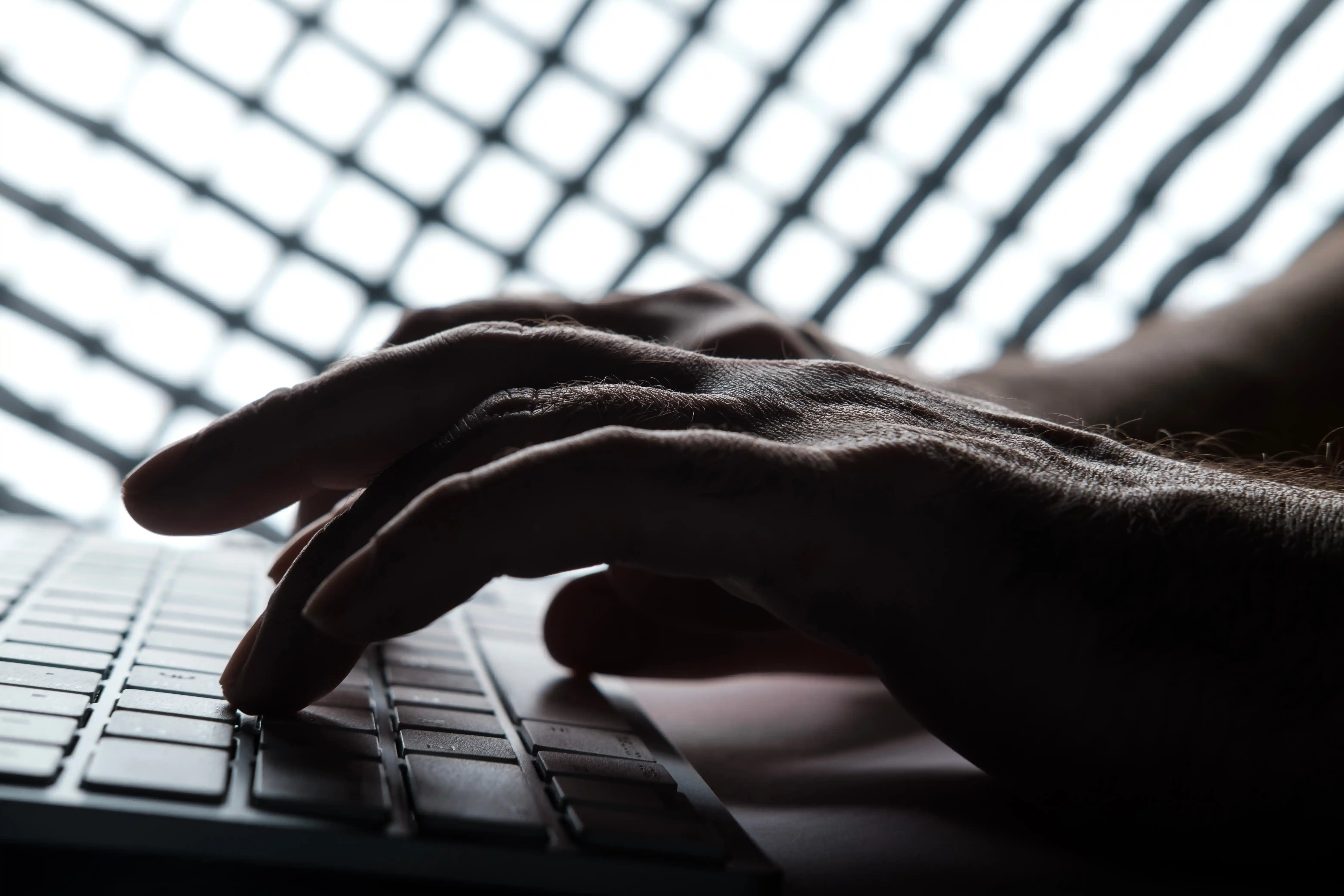 Primer plano de unas manos escribiendo en el teclado de un ordenador portátil, con una cuadrícula luminosa desenfocada al fondo, para ilustrar los riesgos de suplantación de identidad digital.