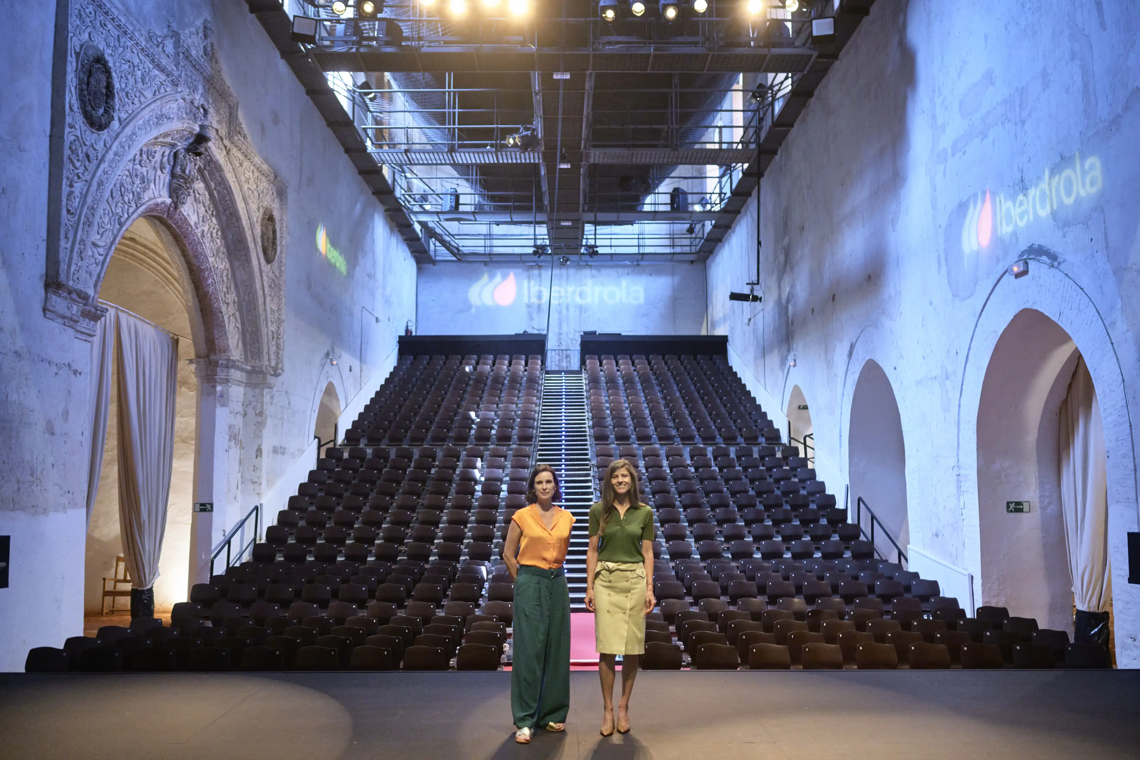 Dos mujeres de pie en escenario de auditorio vacío con proyección de Iberdrola y filas de butacas.