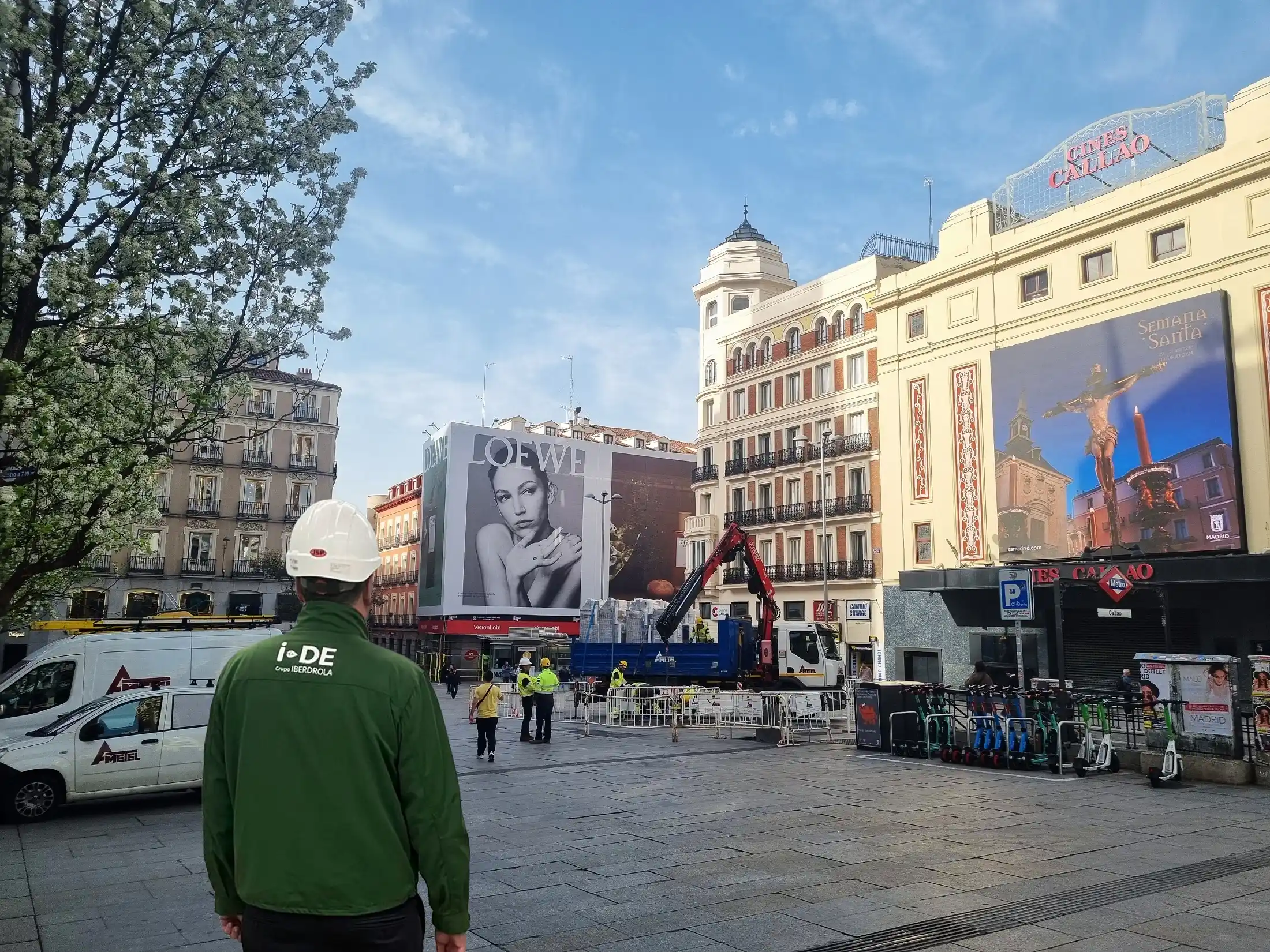 Photograph of a i-DE worker wearing a hard hat in front of a construction site in Plaza Callao, Madrid, with billboards and the Cines Callao building in the background.