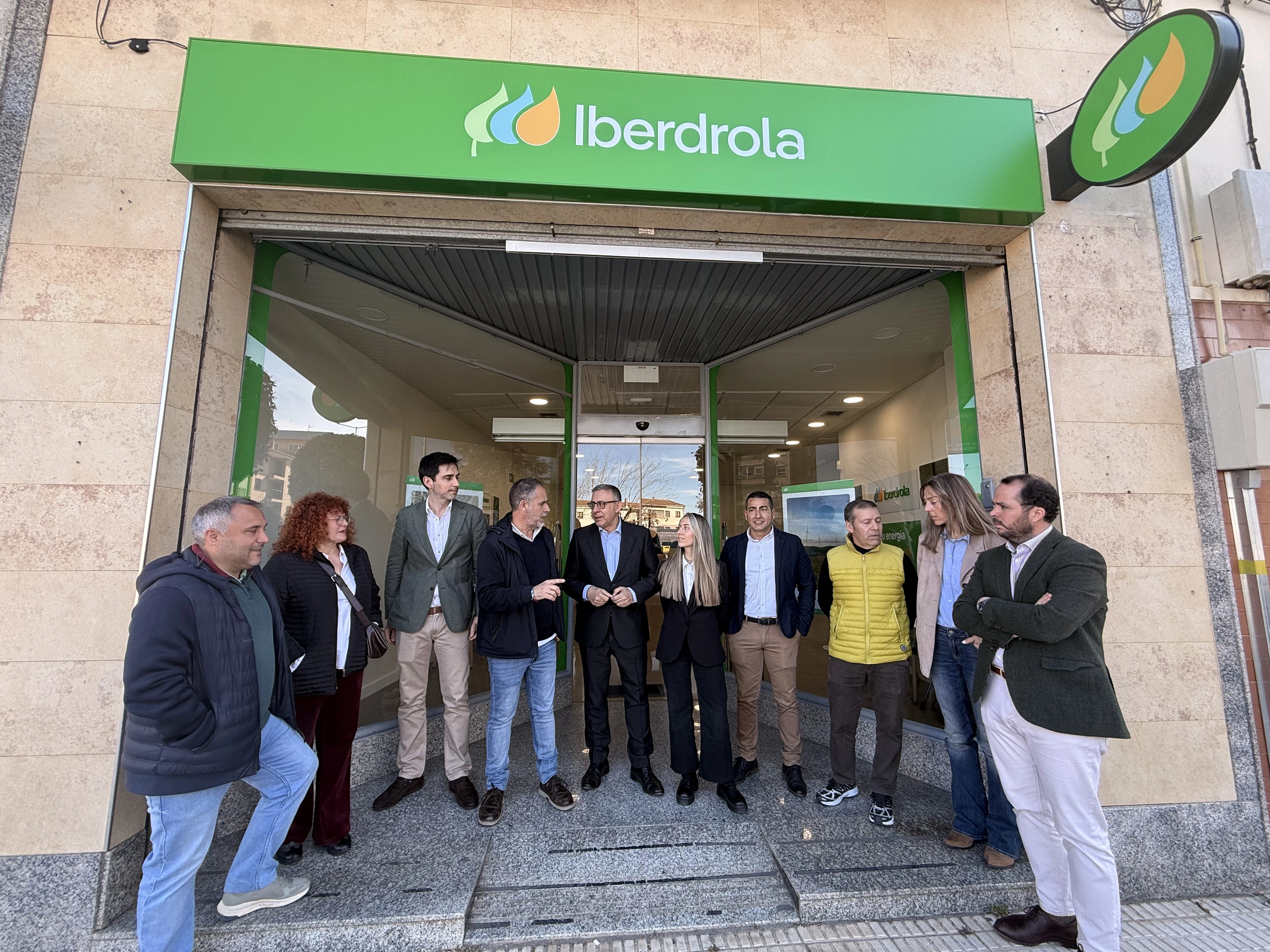 A group of people gathered in front of an Iberdrola store, with the logo visible on the top of the door. The image shows eight people, some standing and others sitting, dressed in casual and professional attire. They are conversing with each other while a man in a suit appears to be speaking to other members of the group.
