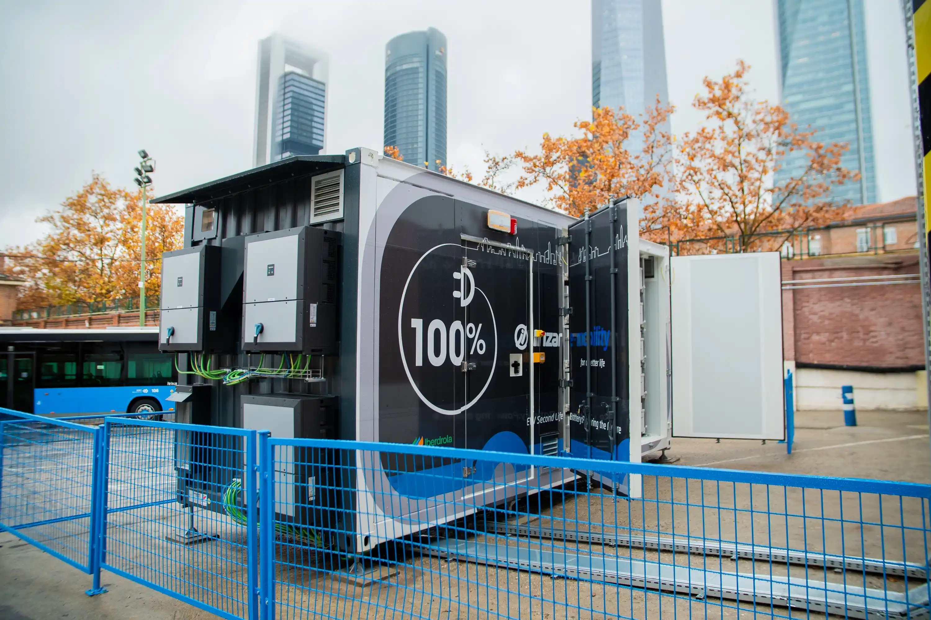 Un contenedor móvil de carga de autobuses eléctricos se encuentra estacionado en una zona al aire libre, con un diseño en colores negro y azul, destacando un símbolo de "100%" en su lado. Está equipado con paneles de energía y cables verdes que conectan a un sistema de carga. En el fondo se observan edificios alto.
