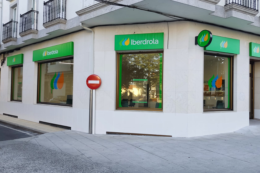 Fachada de una tienda de Iberdrola, ubicada en una esquina. Se observan dos ventanas con el logo de Iberdrola en la parte superior, con una señal de tráfico de 'prohibido el paso' visible al lado.