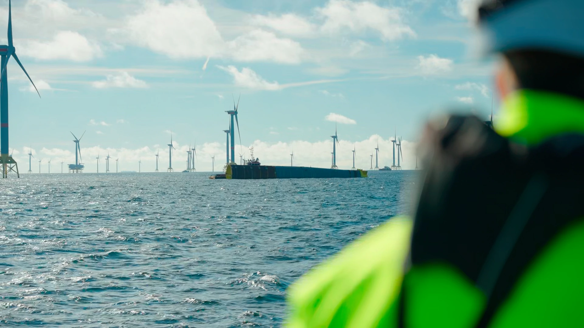 Panoramic view of the ocean with several wind turbines in the background. In the foreground, a person (partially seen from the back) wearing a yellow life jacket is looking towards the sea and turbines. In the center, there is a floating structure with a boat near it.