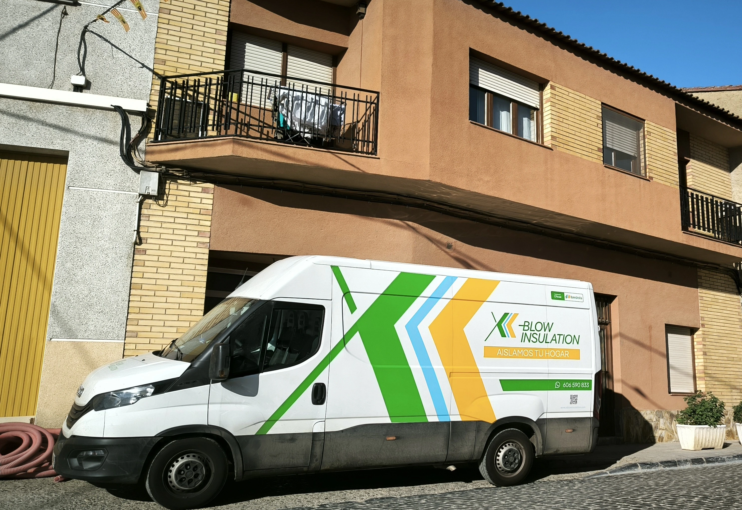 Image of a white van parked in front of a residential building. The van has the inscription "BLOW INSULATION" and the slogan "AISLAMOS TU HOGAR" in green, yellow, and blue.