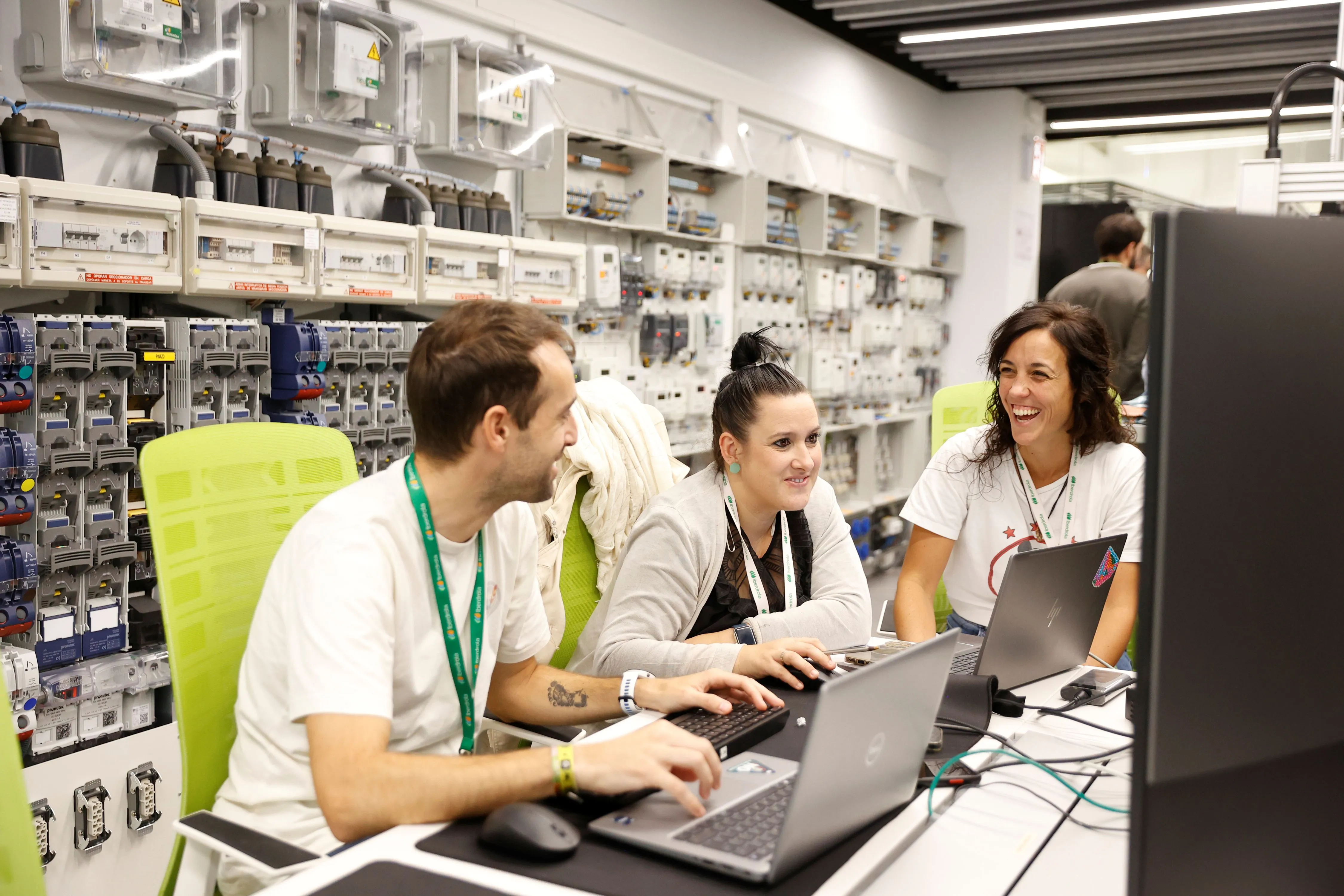 Tres personas trabajan en computadoras portátiles en un espacio de oficina, rodeados de equipos eléctricos y cables, sonriendo mientras colaboran.
