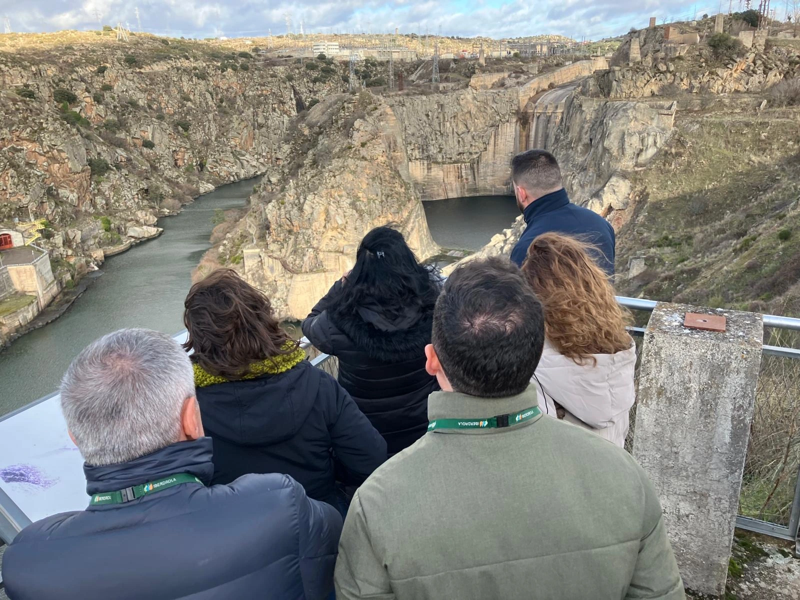 Grupo de personas observando un paisaje rocoso y una instalación hidráulica desde un mirador. Están de espaldas a la cámara, y la escena muestra un desfiladero rodeado de montañas y vegetación. La gente lleva abrigos, y algunas personas tienen el logo de Iberdrola visible en su ropa. En el fondo, se observan estructuras y cables de energía.