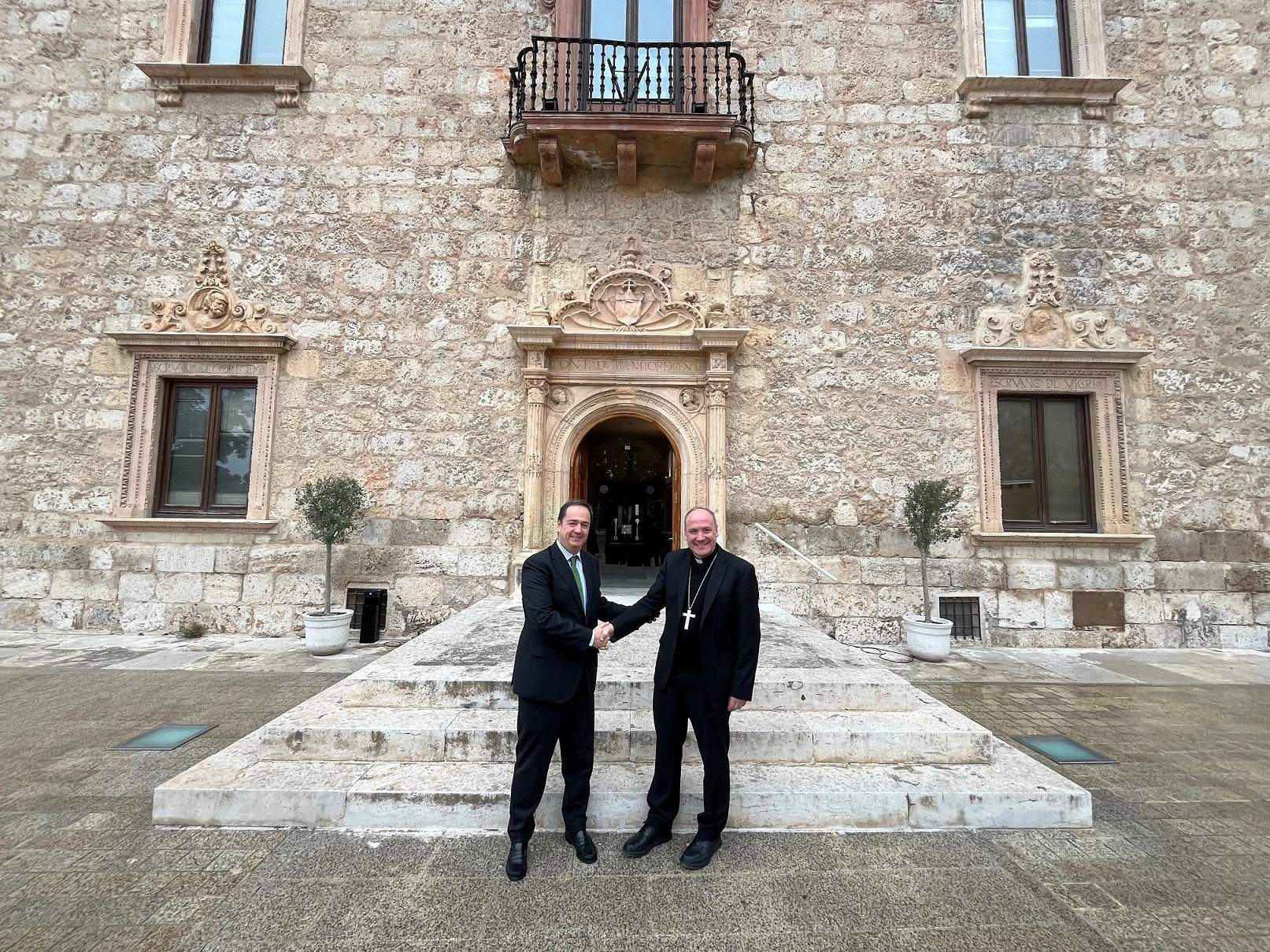 Dos personas se dan la mano frente a una entrada de edificio antiguo. El hombre a la izquierda lleva un traje oscuro con corbata verde, mientras que el hombre a la derecha viste una sotana negra con un crucifijo visible. Ambos están de pie en la escalera de piedra que conduce a la puerta principal del edificio, que tiene una fachada de piedra con detalles arquitectónicos.