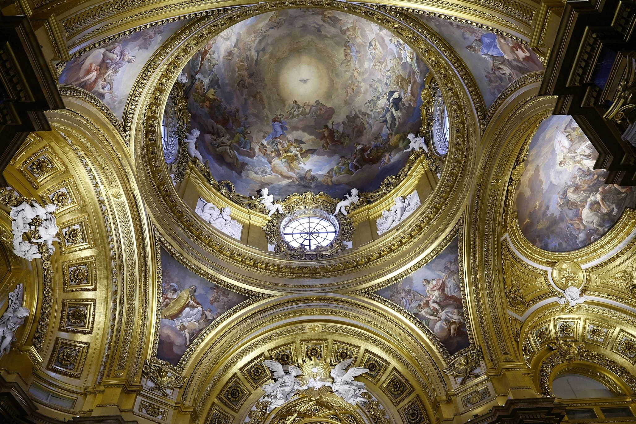 View of the ceiling of the Royal Chapel in the Royal Palace, featuring intricate golden decorations. The ceiling has ornamental details such as moldings and sculpted figures, while the painting in the central dome depicts religious scenes, with a central figure representing a deity surrounded by angels and saints. Light enters through a circular window, highlighting the opulence of the golden details.