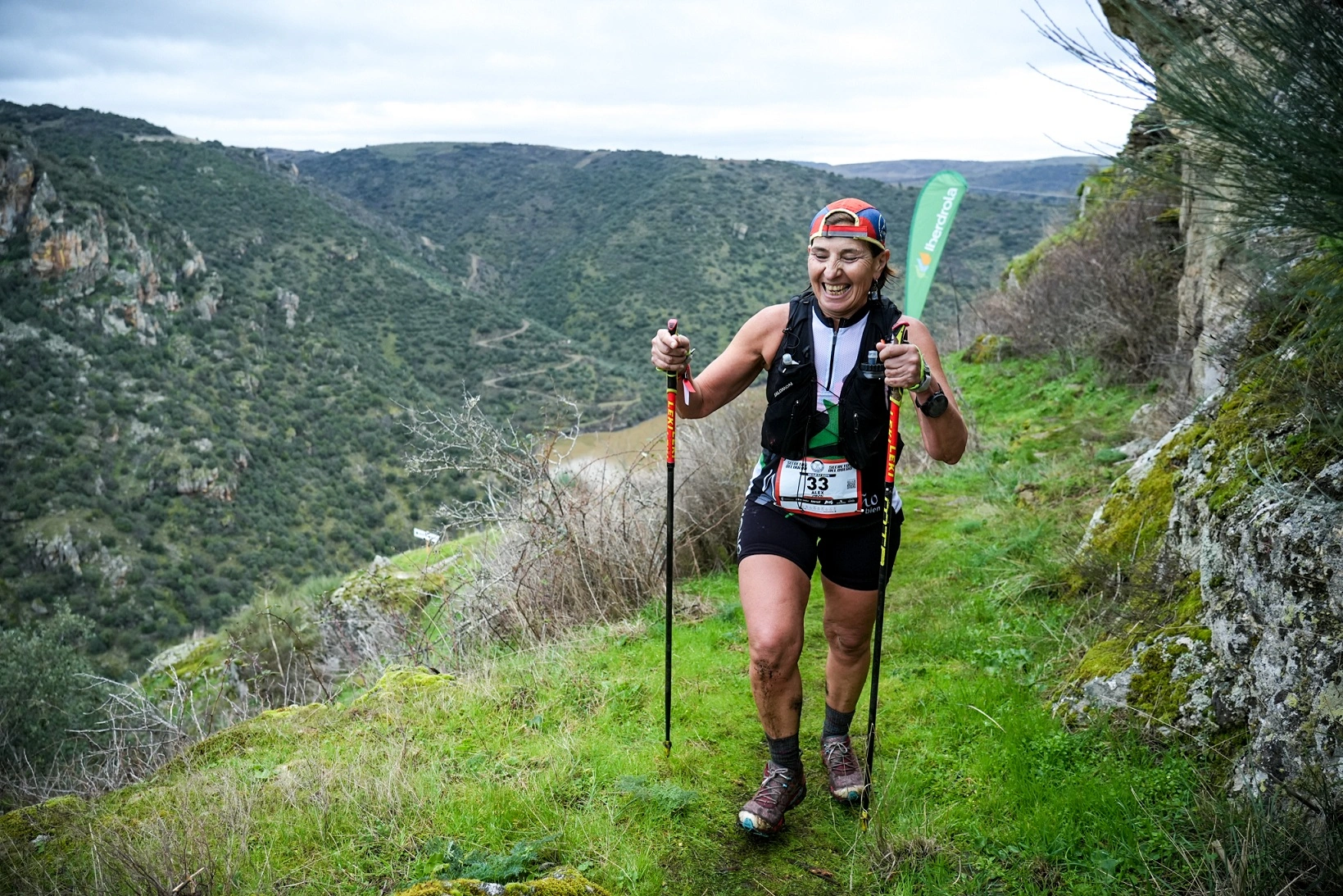 Trail runner moves along a natural path in the Duero canyon, wearing a race bib, with Iberdrola flags in the background.