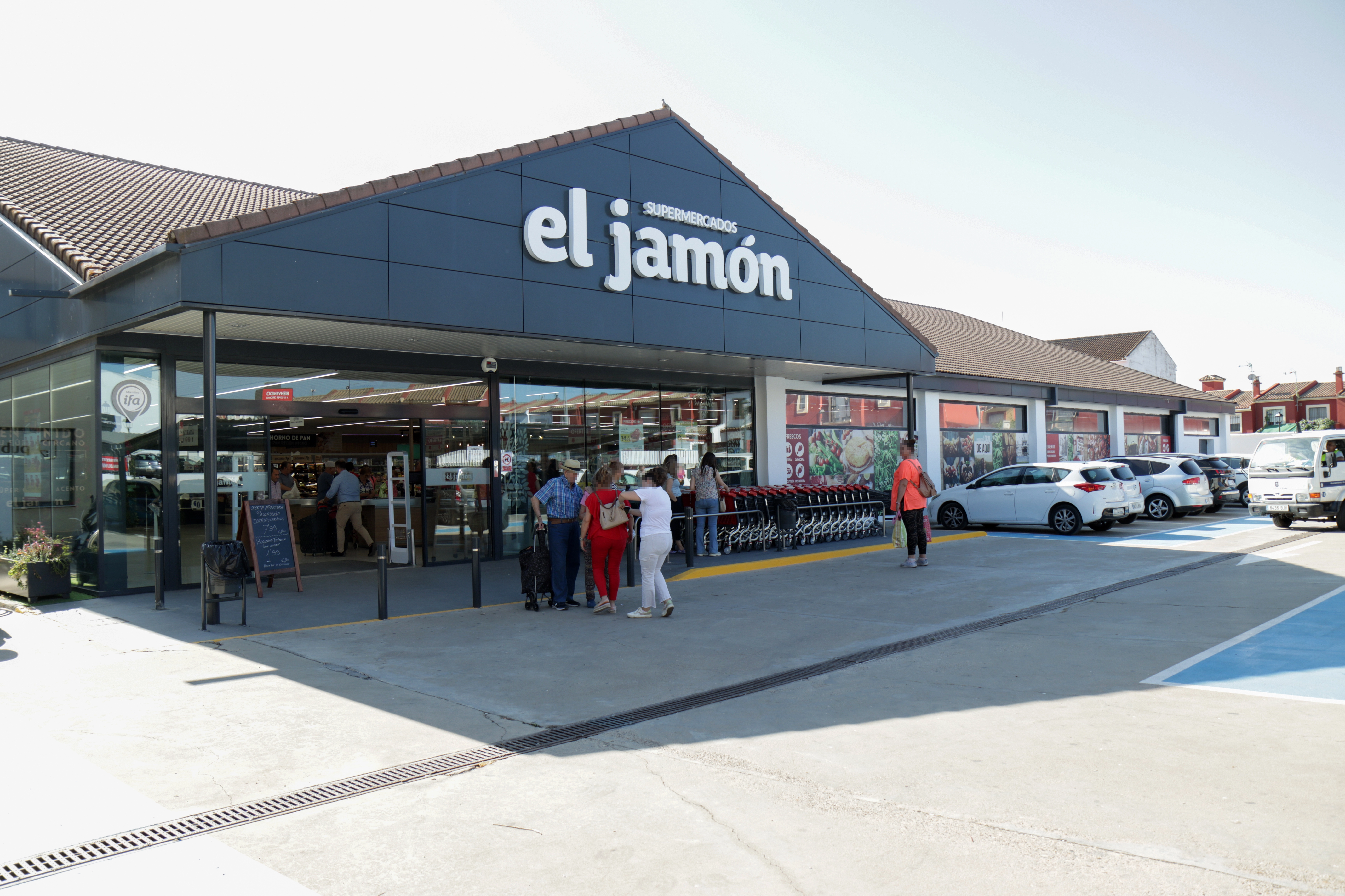 Imagen de la fachada de un supermercado "El Jamón", con el nombre visible en letras grandes sobre el techo. Hay varias personas entrando y saliendo del supermercado, algunas con carritos de compra. En el exterior, se pueden ver vehículos estacionados y un ambiente soleado, con un cielo despejado.