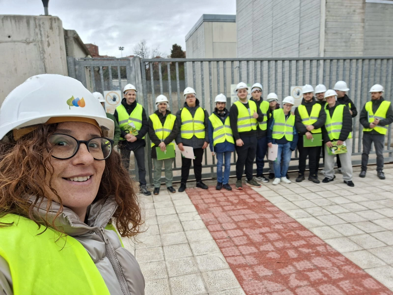 A woman, wearing a safety helmet and reflective vest, poses for a group photo while taking a selfie. She is smiling, and behind her, the group of people also wears safety helmets and reflective vests. They are standing in front of a fence, with a building visible in the background. The woman has the Iberdrola logo on her helmet.