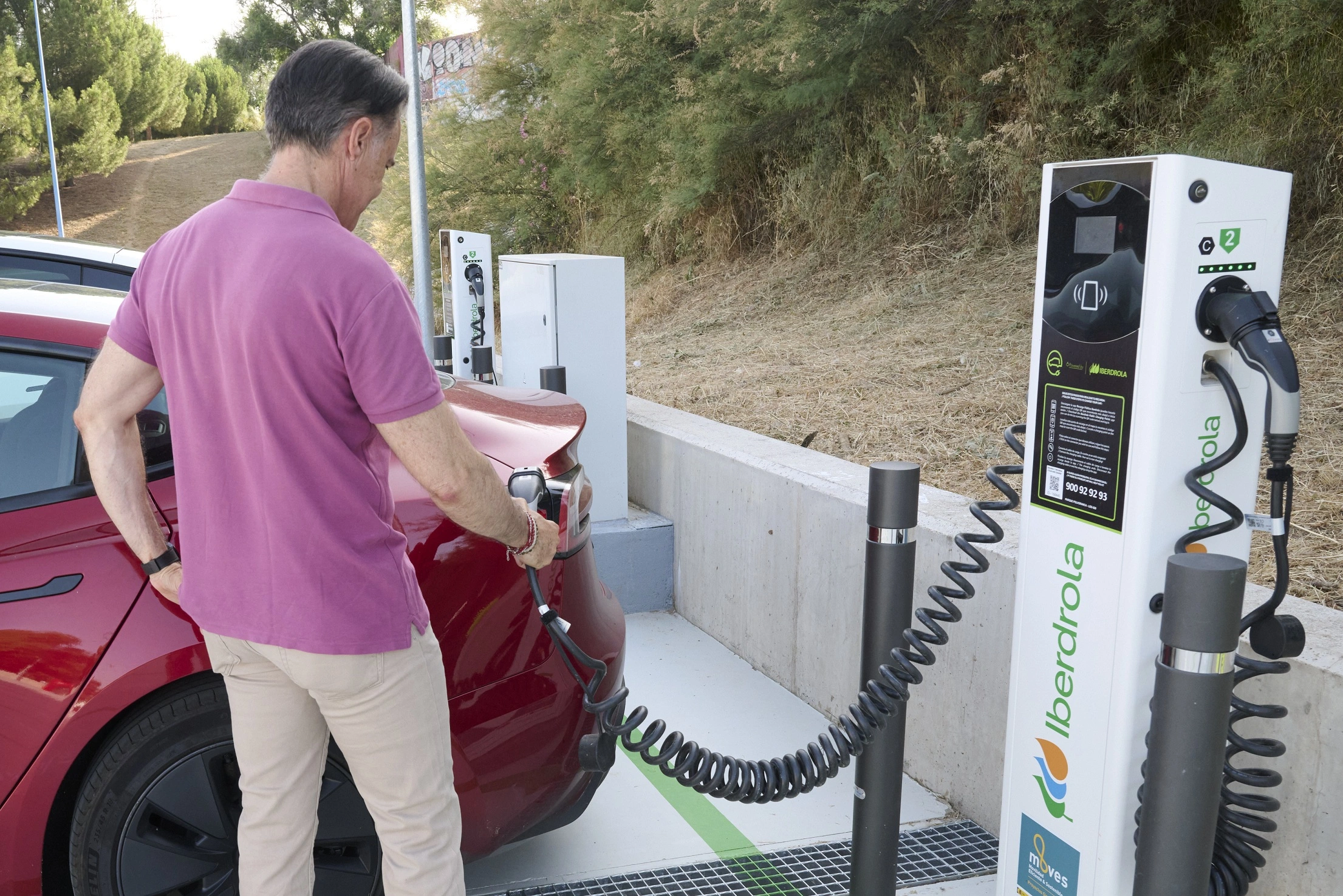 A driver is charging his electric car at an Iberdrola charging point. The charging station is located outdoors, and the Iberdrola logo is clearly visible on the charging station. The driver, with his back turned, holds the charging cable and connects it to the red car. The background shows vegetation and a simple concrete structure.