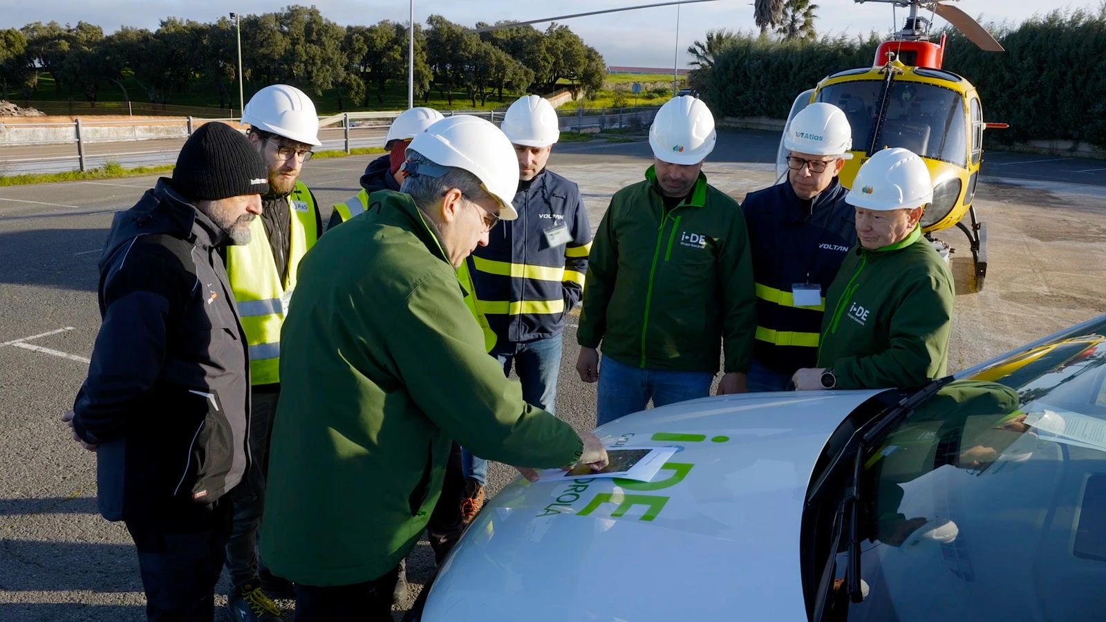 Un grupo de personas, todas con cascos de seguridad y chalecos reflectantes, están reunidas alrededor de un vehículo. Un hombre está usando una tableta para interactuar con la información en el vehículo, mientras los demás observan. Al fondo, se ve un helicóptero amarillo y verde estacionado. El grupo parece estar en una actividad de formación o inspección en un entorno industrial.