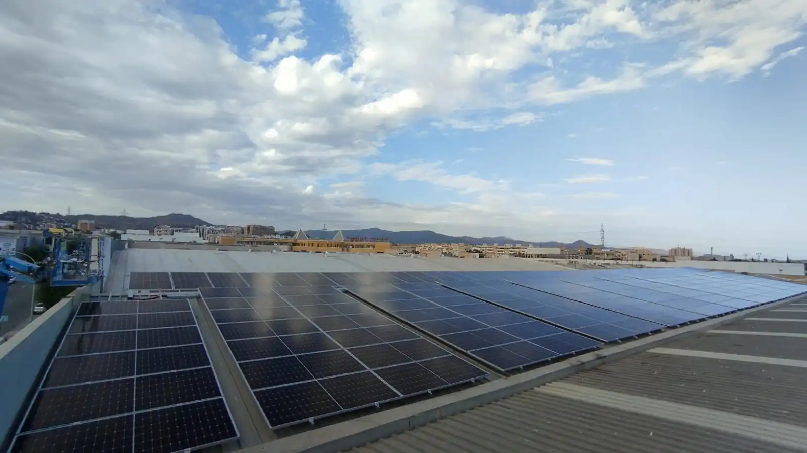 View of a solar panel installation on the roof of a building. The image shows several rows of solar panels covering much of the roof. In the background, there is a partially cloudy sky with some mountains in the distance.