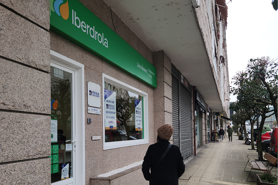 View of an Iberdrola store on a street, with a green sign on the facade displaying the company's logo. In the foreground, a person walks toward the store, wearing a coat and a hat. In the background, the pedestrian walkway of the street is visible with trees and benches. The store has windows with visible advertisements, and a closed shop is next to it.
