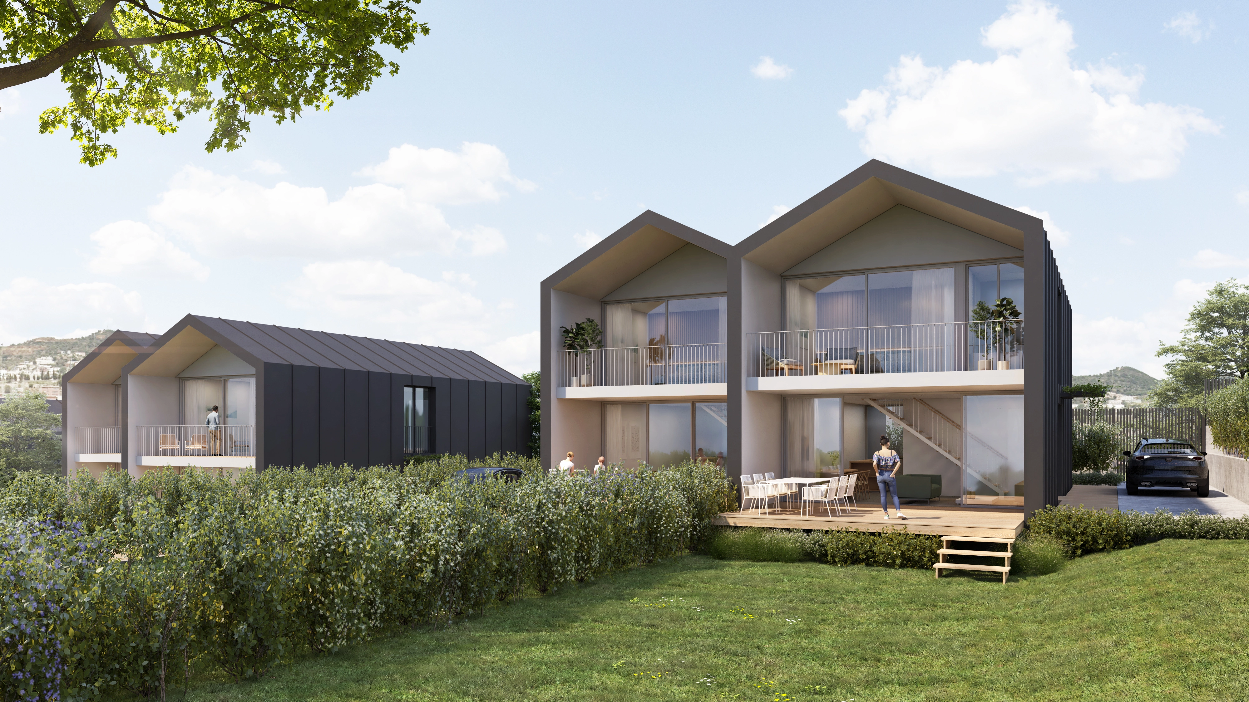 Vista exterior de dos viviendas unifamiliares modernas de dos plantas con grandes ventanales y balcones. Las casas tienen fachadas en tonos claros y oscuros, con diseño contemporáneo y techos a dos aguas. En primer plano se ve un jardín con césped y setos, así como una terraza de madera con mesa y sillas. Hay personas en el exterior y un coche aparcado junto a una de las viviendas, en un entorno residencial tranquilo.