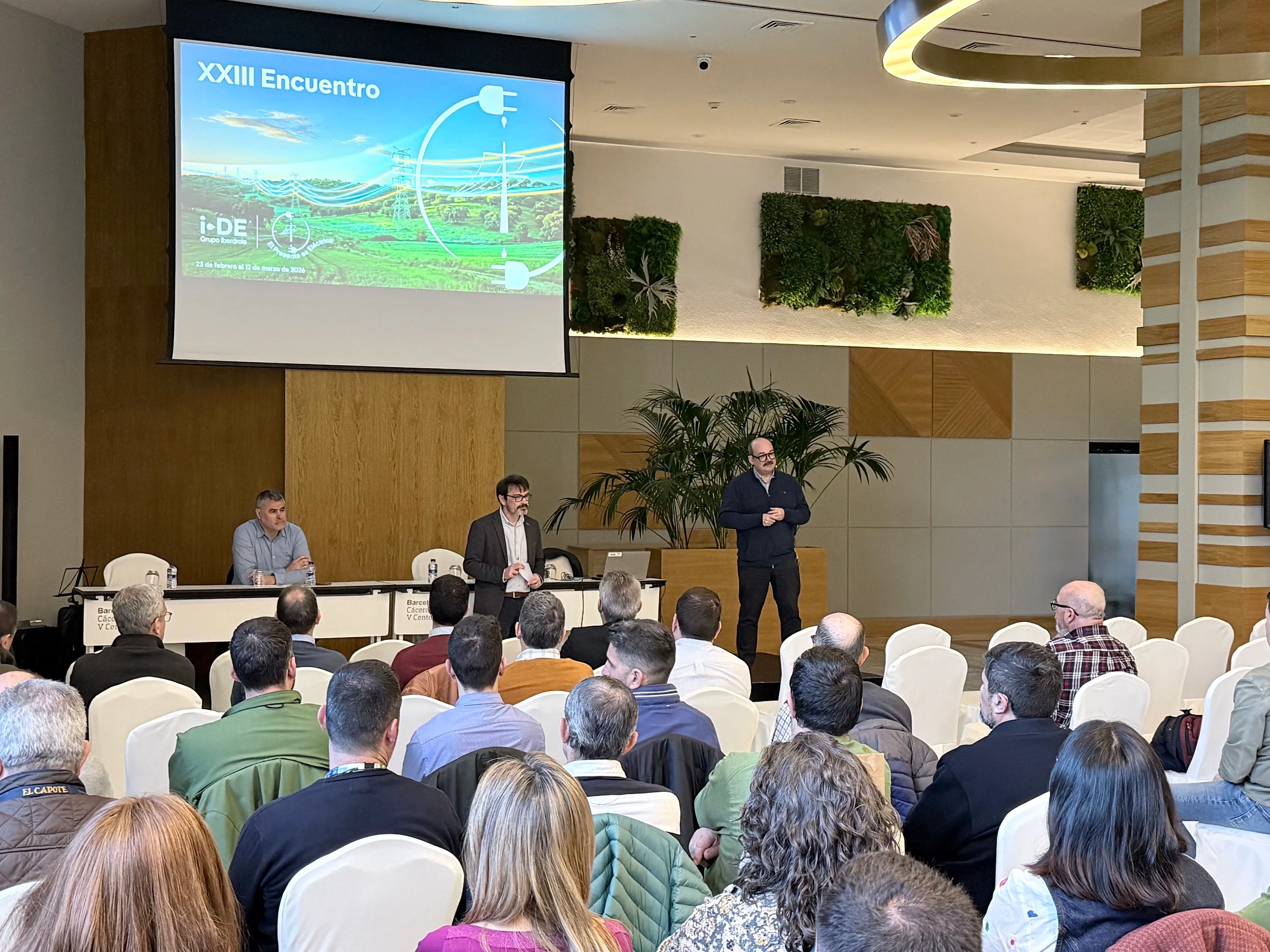 Conference room with an audience attending a presentation. On stage, three men are taking part in a panel discussion: two seated at a table and one standing while addressing the audience. The projected screen reads “XXIII Encuentro” alongside the i-DE Grupo Iberdrola logo and an image related to power networks. The room features modern decor and green wall panels.