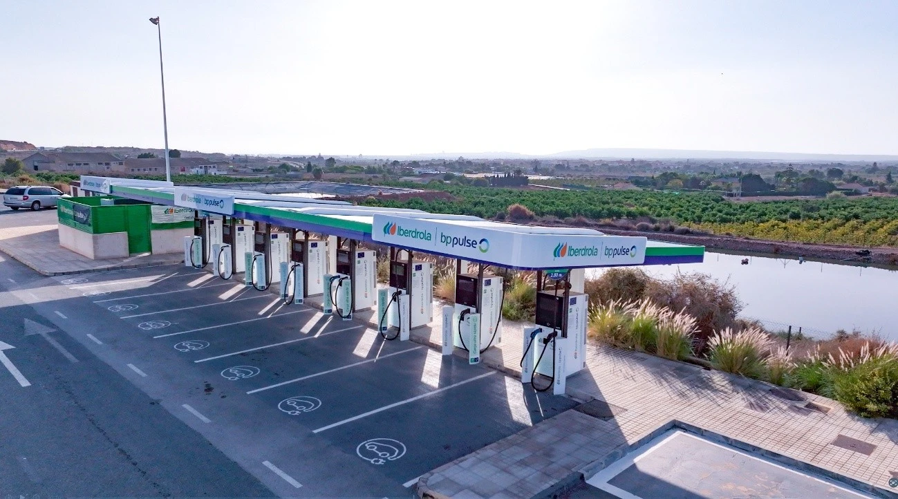 Vista aérea de una estación de carga de vehículos eléctricos de Iberdrola y BP Pulse. Se pueden ver varias estaciones de carga dispuestas en línea, con el logo de ambas empresas en el techo de cada estación. En el fondo, se observa un paisaje rural con campos y montañas a lo lejos. El área está despejada y bien organizada, con espacios de estacionamiento para vehículos eléctricos.