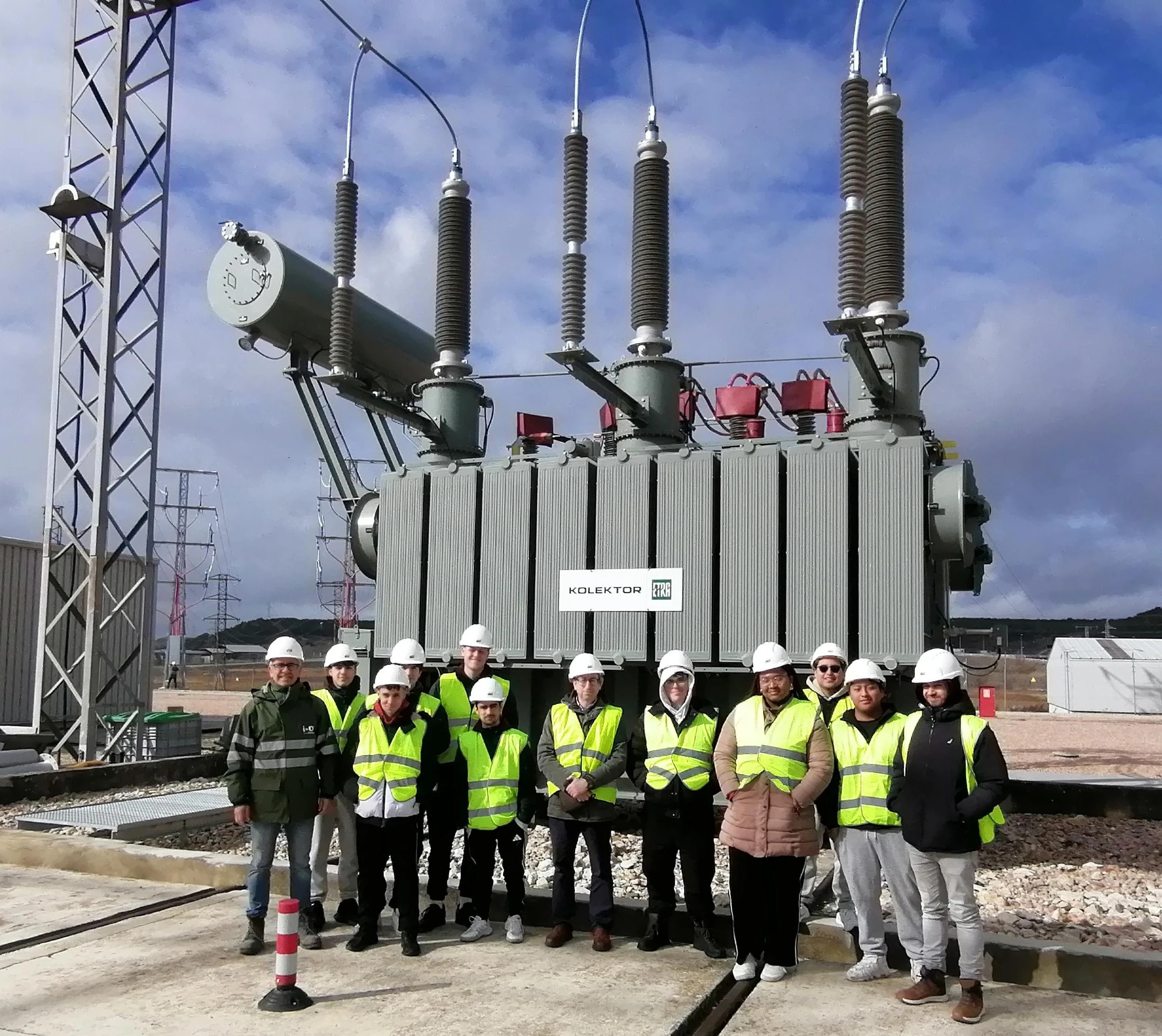 A group of people is posing in front of a transformer at an electrical substation. They are all wearing safety helmets and yellow reflective vests. In the background, the infrastructure of the substation is visible, with cables and electrical transmission towers. The group appears to be on an educational or professional visit, in an industrial and open environment.