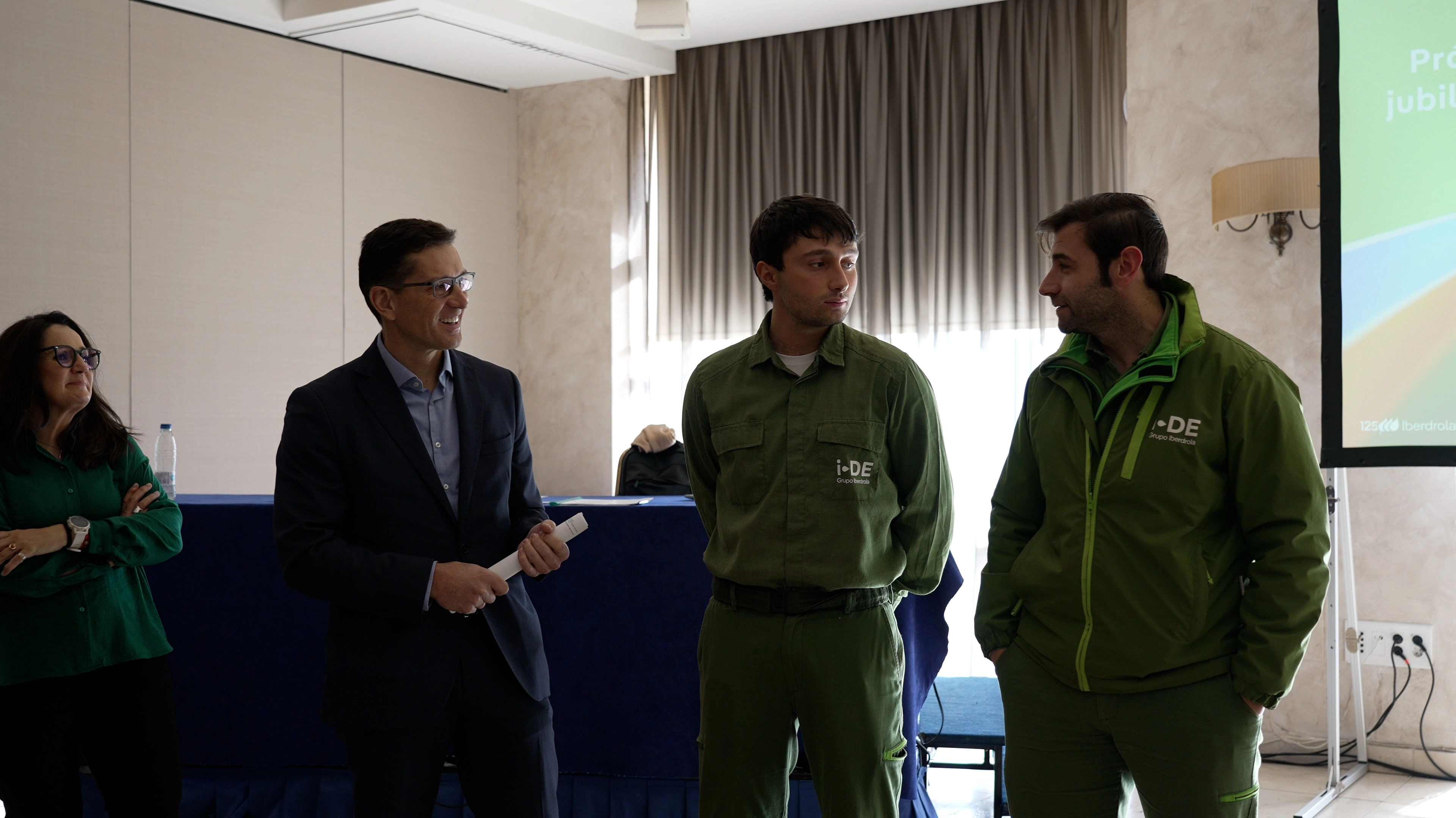 Un grupo de personas está en una sala durante un evento o presentación. De izquierda a derecha, una mujer con una camisa verde sonríe y observa a los dos hombres que están de pie. Los hombres llevan chaquetas verdes con el logo de i-DE y parecen estar en una conversación o charla. En el fondo, se ve una pantalla con el logo de Iberdrola. El ambiente es de un encuentro o evento corporativo.