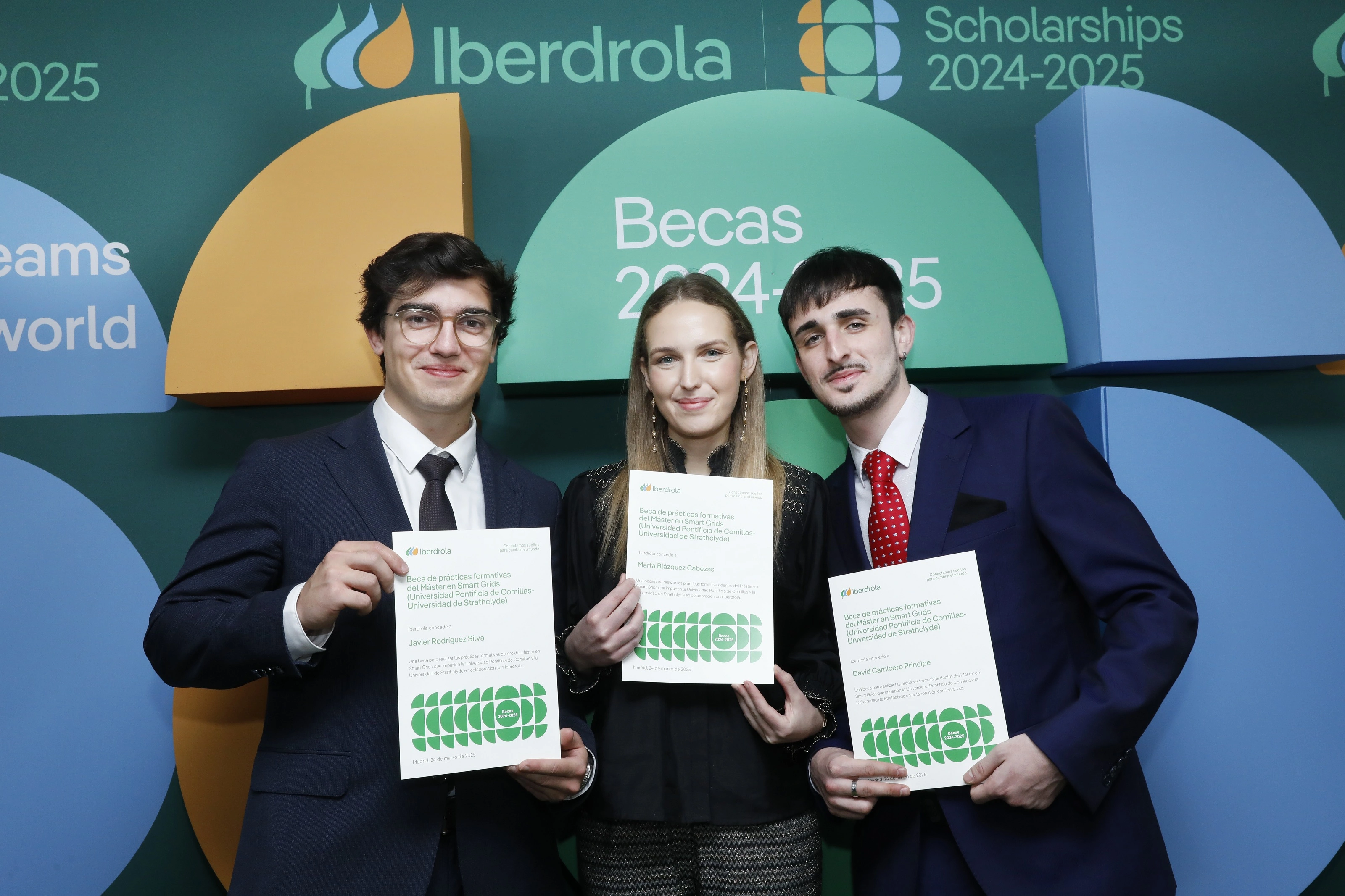 Tres jóvenes posan frente a un fondo corporativo de Iberdrola que anuncia las Becas 2024-2025. Cada uno sostiene un diploma de la beca, vestidos con traje formal. En el fondo se leen los textos “Iberdrola” y “Scholarships 2024-2025” junto a elementos gráficos en verde, azul y amarillo.