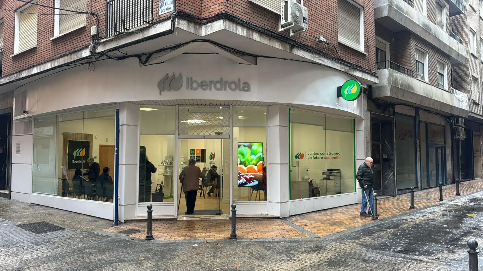 Vista exterior de una tienda de Iberdrola en una calle de ciudad. La fachada muestra el logo de Iberdrola en color verde y blanco, junto con el eslogan "Juntos construimos un futuro sostenible" en la ventana. Un hombre mayor camina por la acera cerca de la entrada, mientras dentro de la tienda se pueden ver algunas personas sentadas. El día está nublado y la calle está mojada, lo que sugiere que ha llovido recientemente.