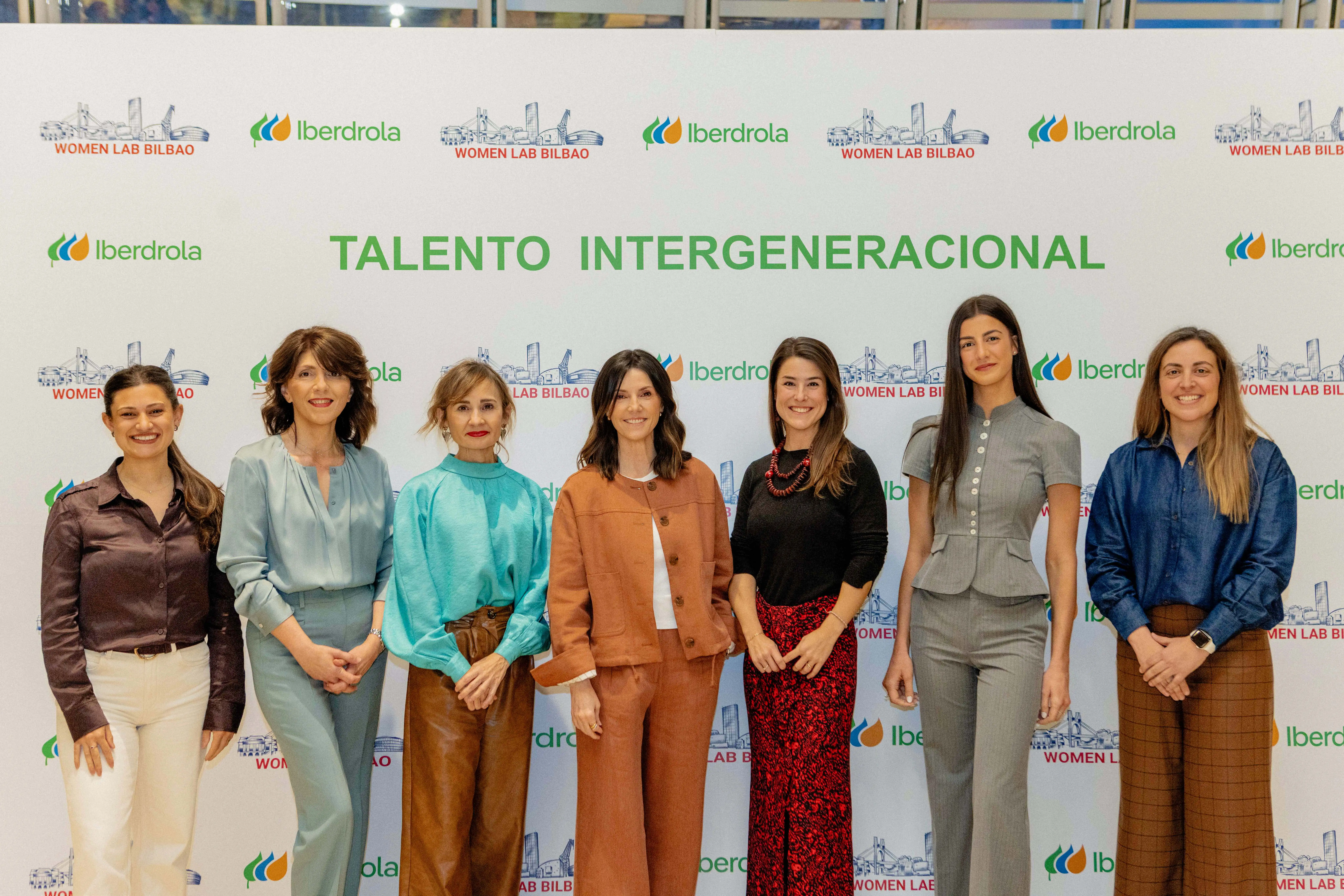 A group of women is posing in front of a sign with the Iberdrola logo and the slogan "Intergenerational Talent" at the Women Lab Bilbao event. Each woman is wearing modern, colorful attire and they all look happy. The image conveys a sense of collaboration and female empowerment in a professional setting.
