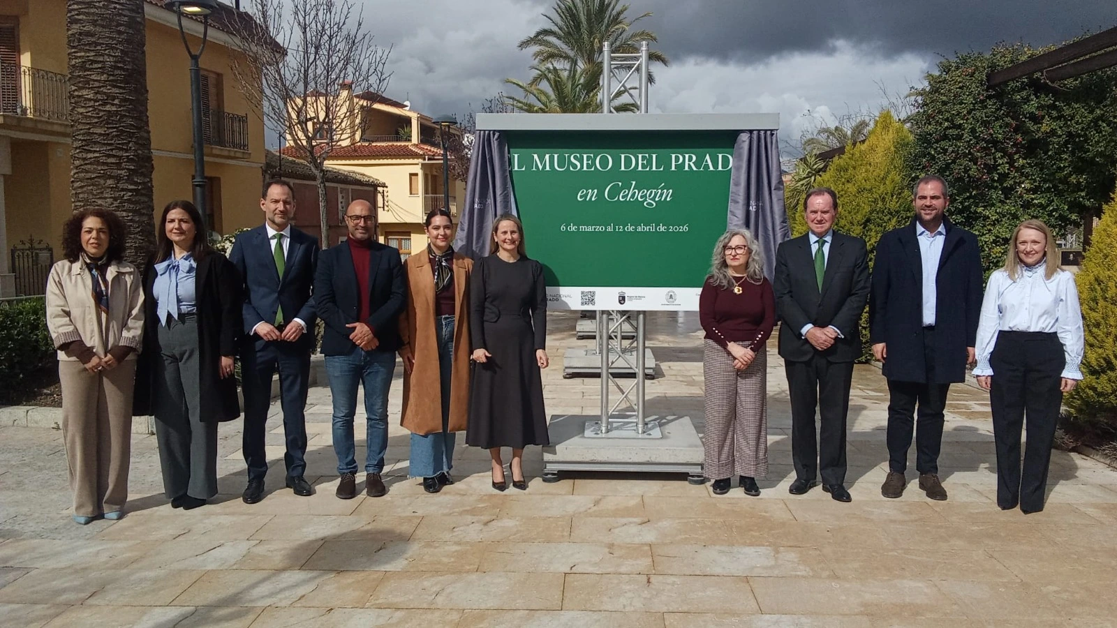 A group of people stands in front of a sign announcing the Museo del Prado in Cehegín exhibition. The group consists of various individuals dressed formally, wearing suits and coats, standing in an outdoor square. In the background, there are buildings and an urban landscape. The sign highlights the event's dates, running from March 6 to April 12, 2026.