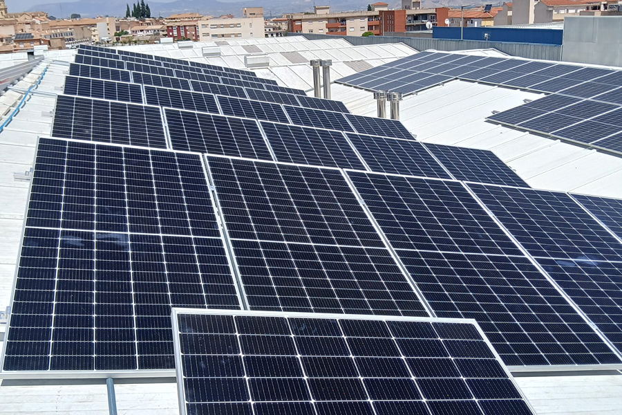 Aerial view of a roof with solar panels installed in a community in Granada. The panels cover a large portion of the roof, capturing sunlight to generate renewable energy. In the background, the buildings of the city can be seen with a clear sky line.
