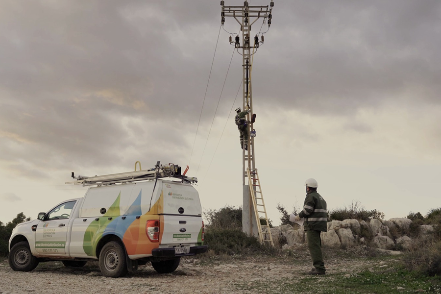 Un técnico trabaja en lo alto de un poste eléctrico mientras otro compañero observa desde el suelo junto a una furgoneta de i-DE Grupo Iberdrola. El vehículo está estacionado en un entorno rural con rocas y vegetación, y el cielo aparece nublado. La escena muestra labores de mantenimiento en la red eléctrica.