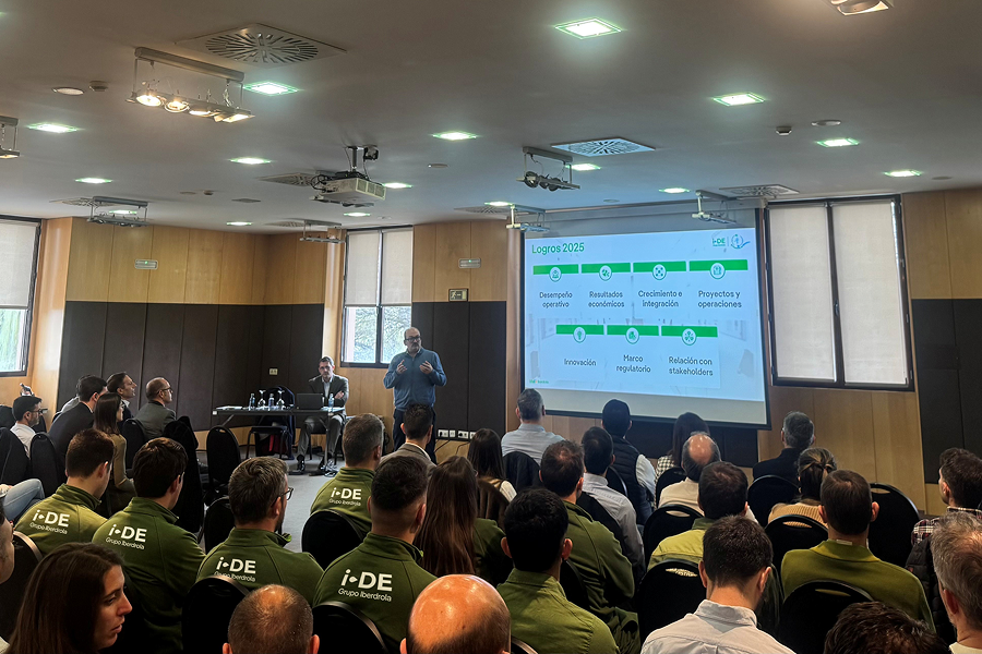 Una sala de conferencias donde varias personas asisten a una presentación corporativa. Un ponente habla frente a una pantalla que muestra una diapositiva titulada “Logros 2025”, mientras el público escucha sentado. Algunos asistentes llevan chaquetas verdes con el logotipo de i-DE Grupo Iberdrola, lo que indica un encuentro profesional de la compañía.