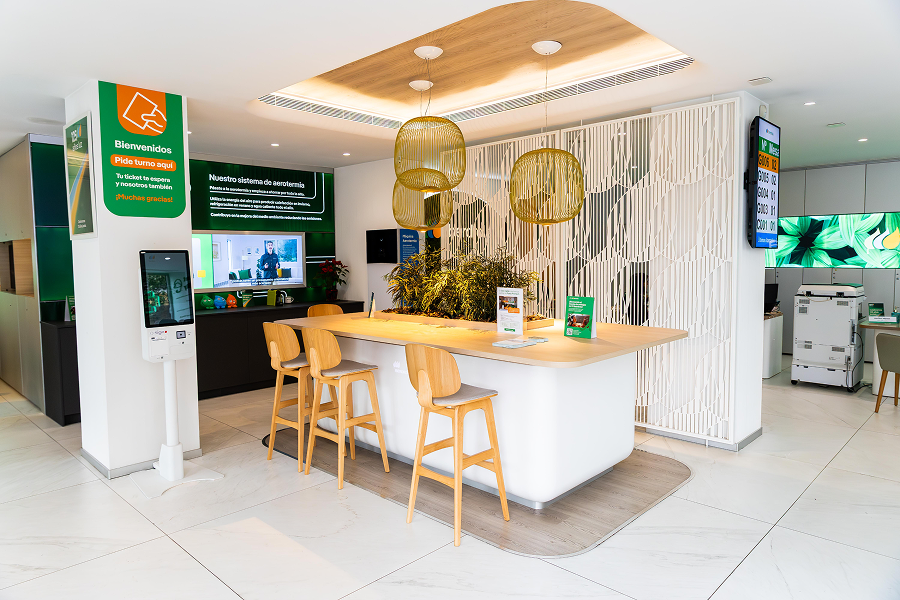 Interior de un punto de atención al cliente de Iberdrola España con diseño moderno y luminoso. En el centro hay una mesa alta de madera con taburetes y plantas decorativas, iluminada por lámparas colgantes. En las paredes se ven pantallas informativas y carteles de bienvenida, además de un terminal digital para atención al cliente.