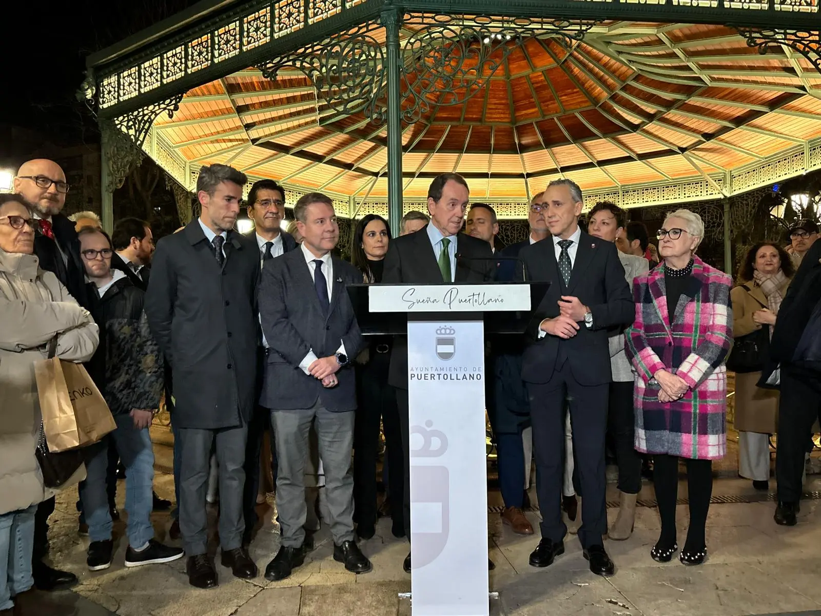 En la imagen, Emiliano García-Page, presidente de la Junta de Comunidades de Castilla-La Mancha, y Jaime Alfonsín, presidente de la Fundación Iberdrola España, se encuentran en un evento al aire libre, en frente de una estructura de hierro con un techo de colores. Ambos están en el podio con micrófono, aparentemente realizando un discurso, mientras otras personas están atentas a la ceremonia. La imagen corresponde a un evento oficial o de celebración en el municipio de Puertollano, con varias personas de pie alrededor, algunas con trajes formales.
