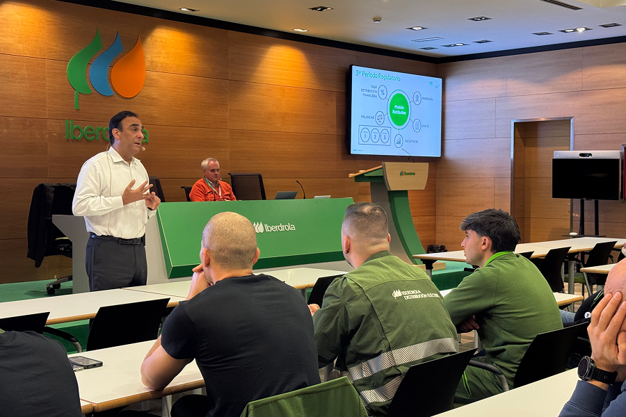 Un hombre está dando una presentación en una sala de reuniones de Iberdrola. En el fondo se puede ver una pantalla con un gráfico relacionado con el "3º Período Regulador". En primer plano, varios empleados de Iberdrola, vestidos con ropa de trabajo verde, están prestando atención a la presentación.