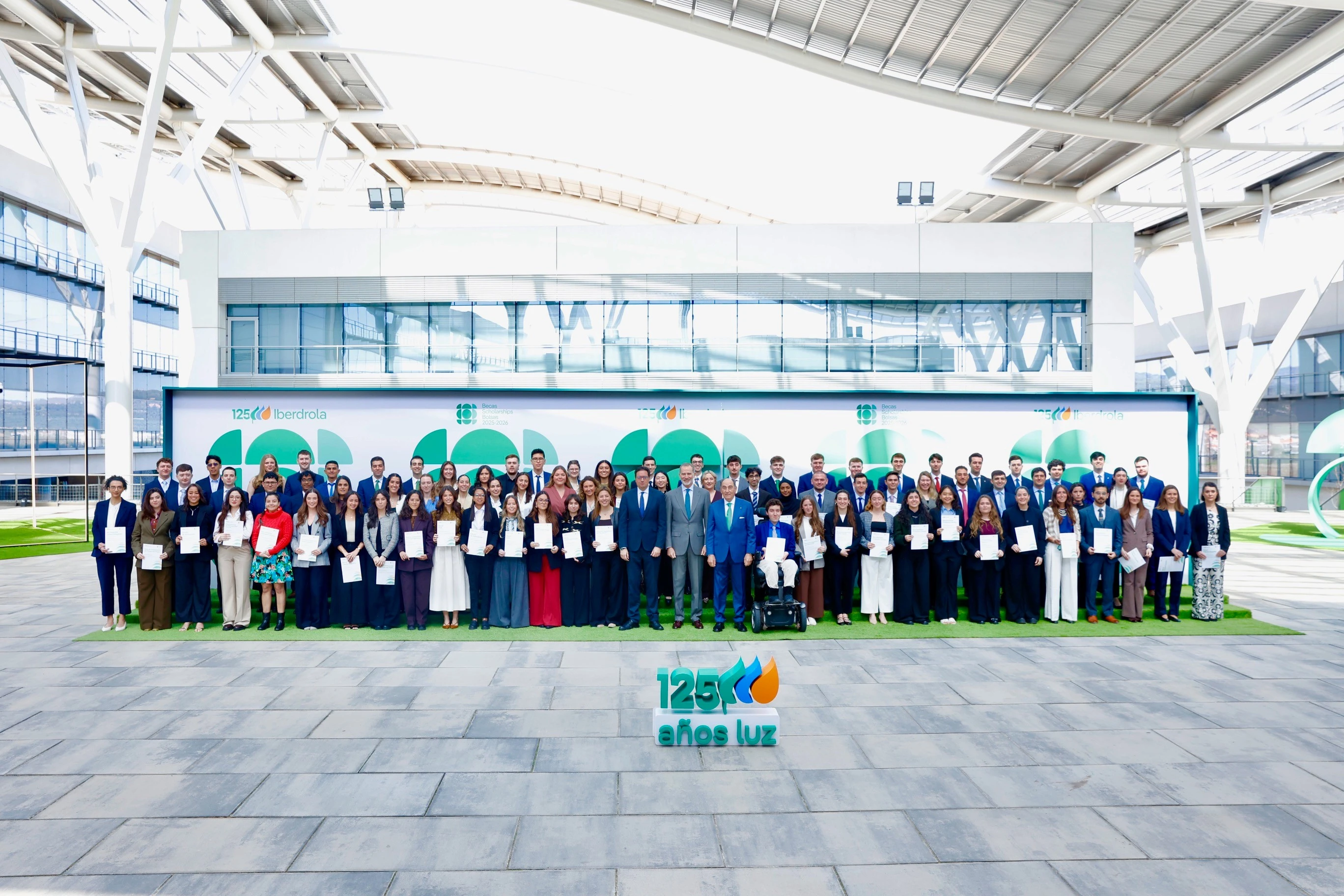 Grupo de personas posando con certificados frente a un cartel con el logo de Iberdrola, en un evento de becas. Entre los asistentes están Ignacio Galán, presidente del Grupo Iberdrola, y el rey Felipe VI, ambos vestidos de manera formal. Todos los participantes están sonrientes para la foto.