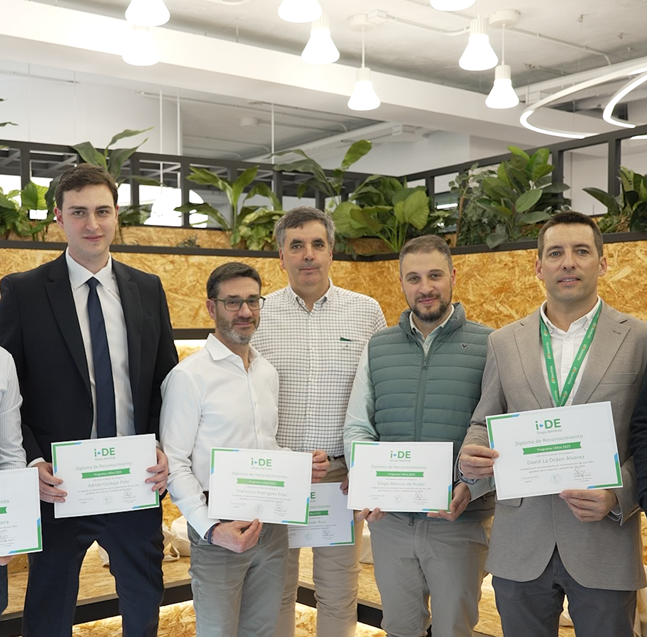 Group of people posing with recognition diplomas during the i-DEas 2026 Awards ceremony, in a modern environment with decorative plants.