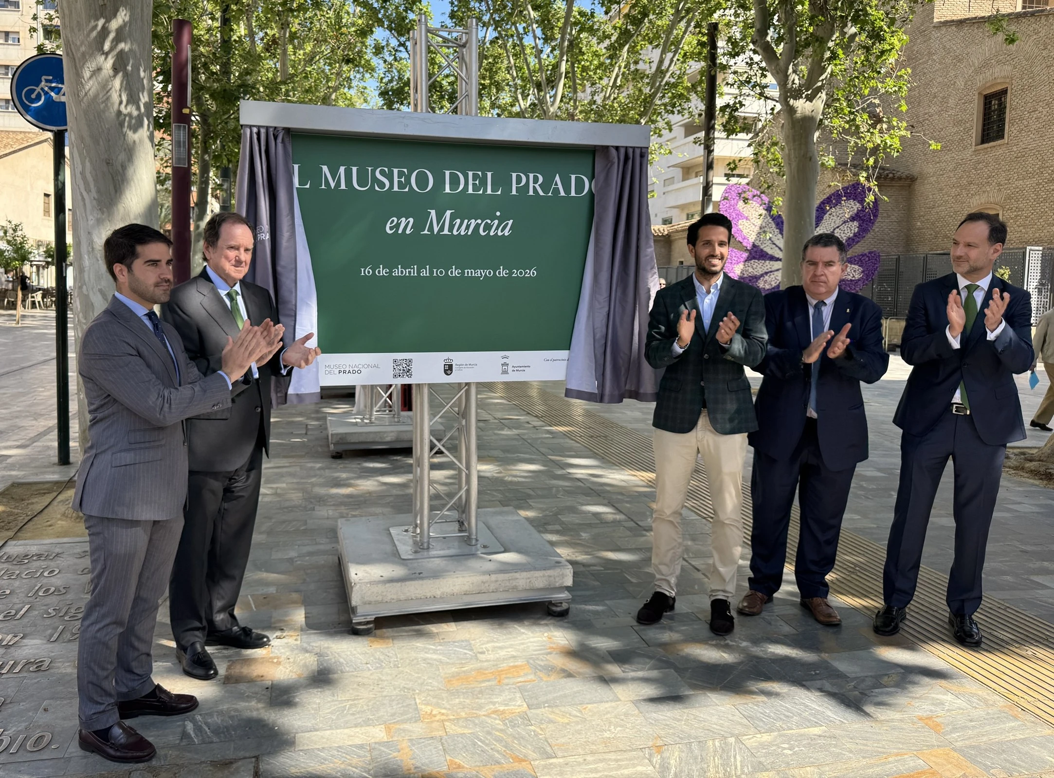 Inauguration of the "El Museo del Prado en Murcia" sign, with representatives clapping and the sign covered, at an outdoor event.