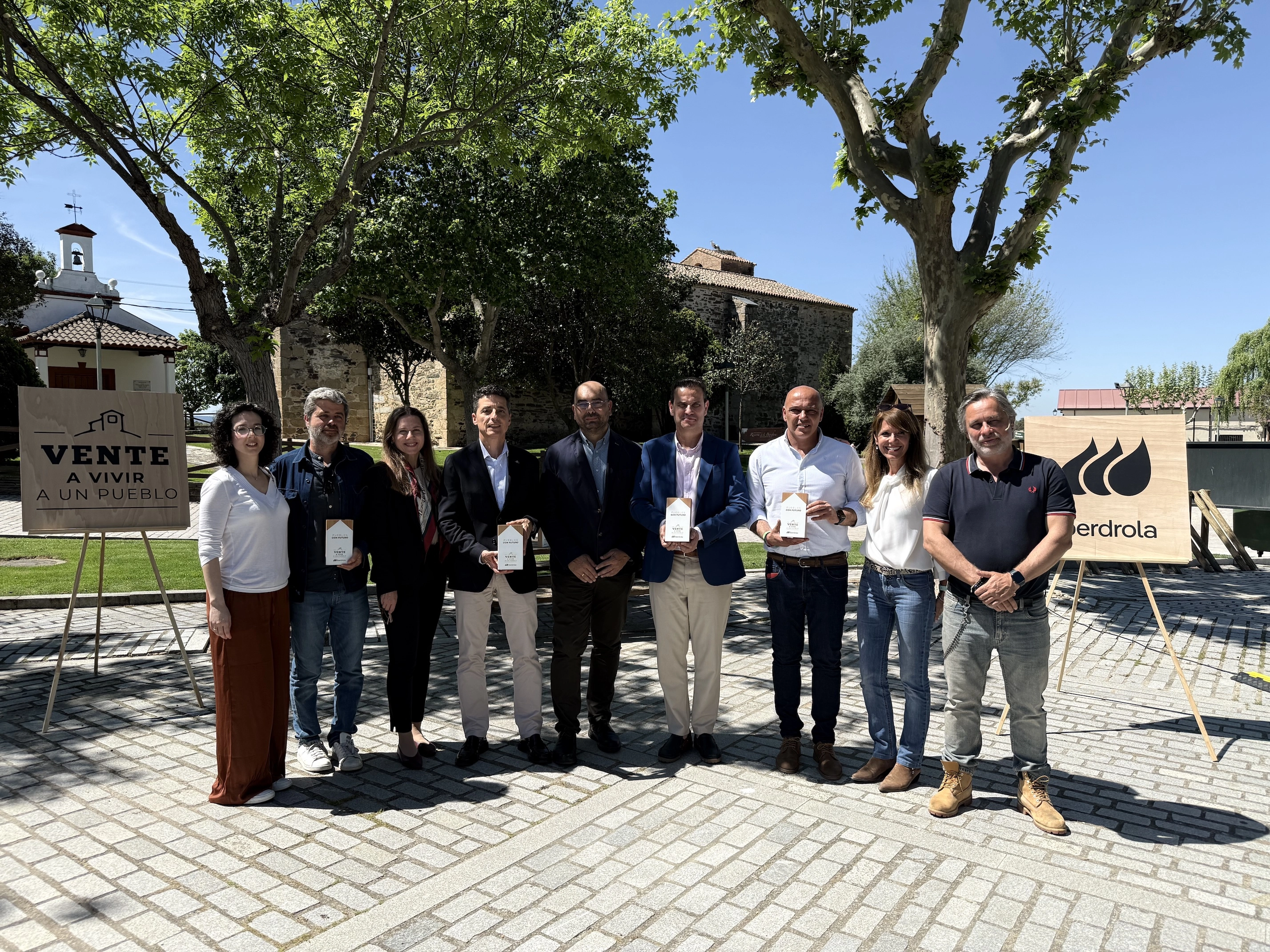 Group of people posing with awards at an outdoor ceremony, with Iberdrola and "Vente a vivir a un pueblo" campaign signs in the background.