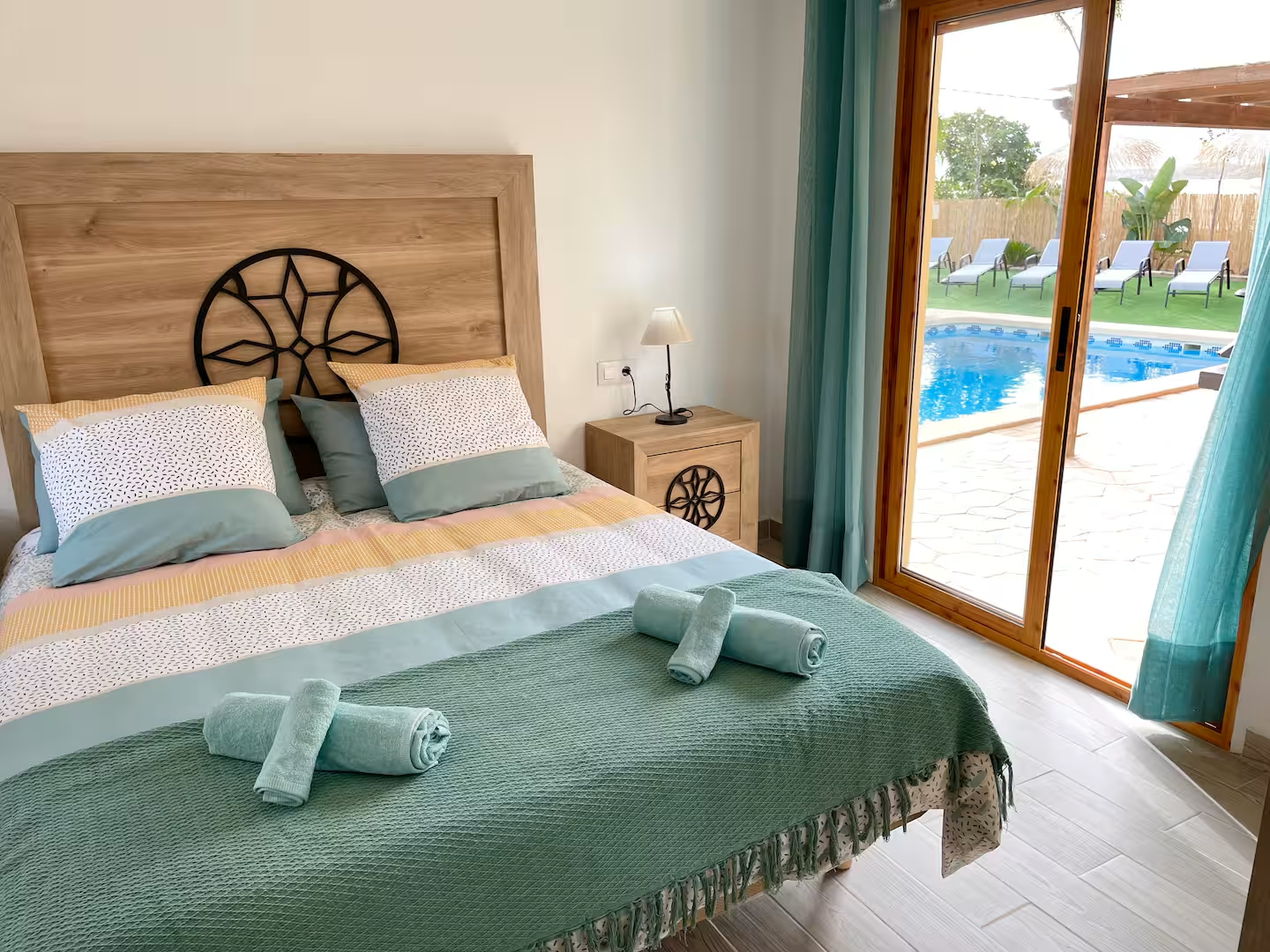 Habitación en casa rural con vistas a la piscina, cama decorada con colcha y toallas, con luz natural entrando por la ventana.