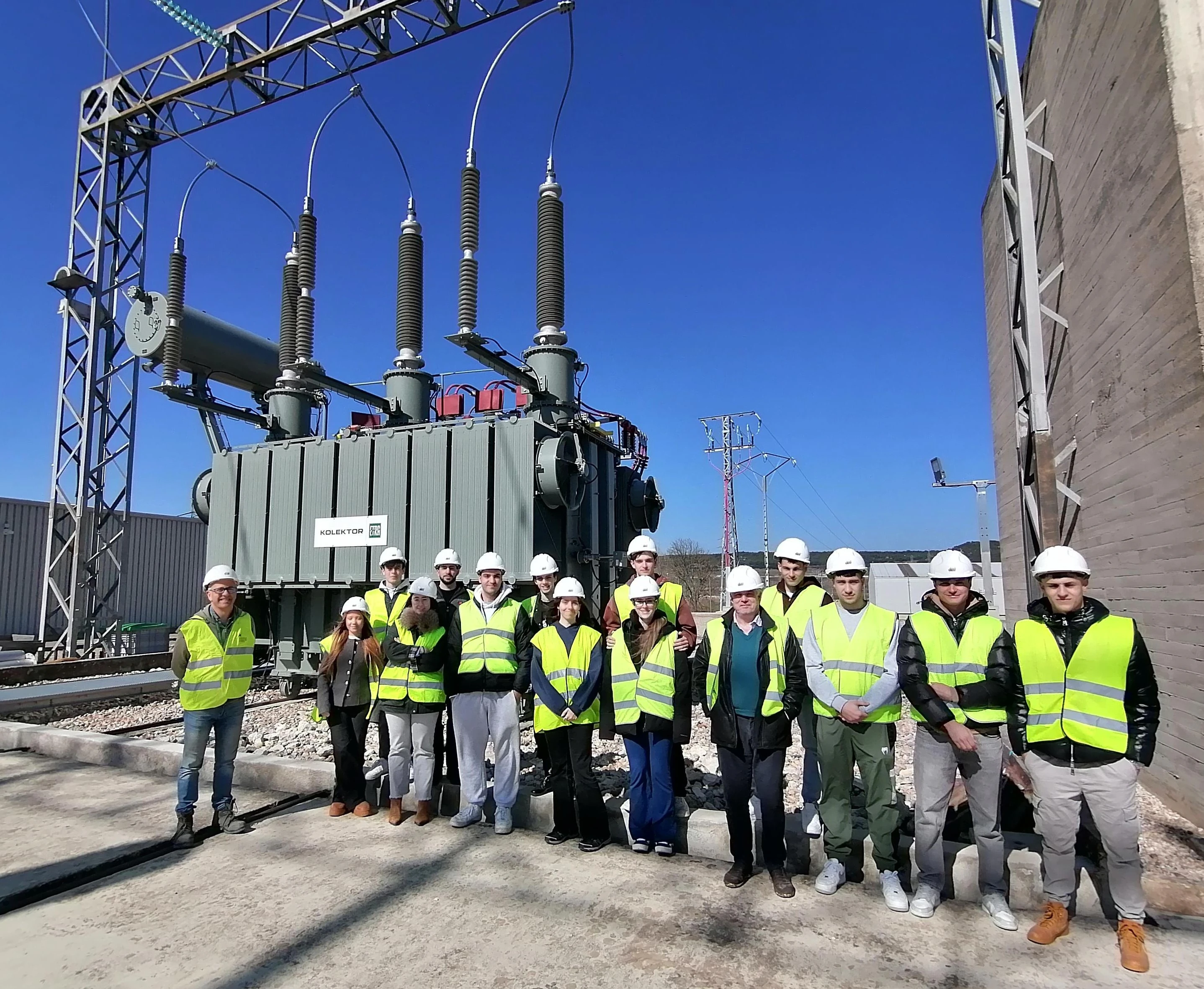 Group of students posing in front of an electrical transformer at a substation, wearing safety equipment like helmets and reflective vests.
