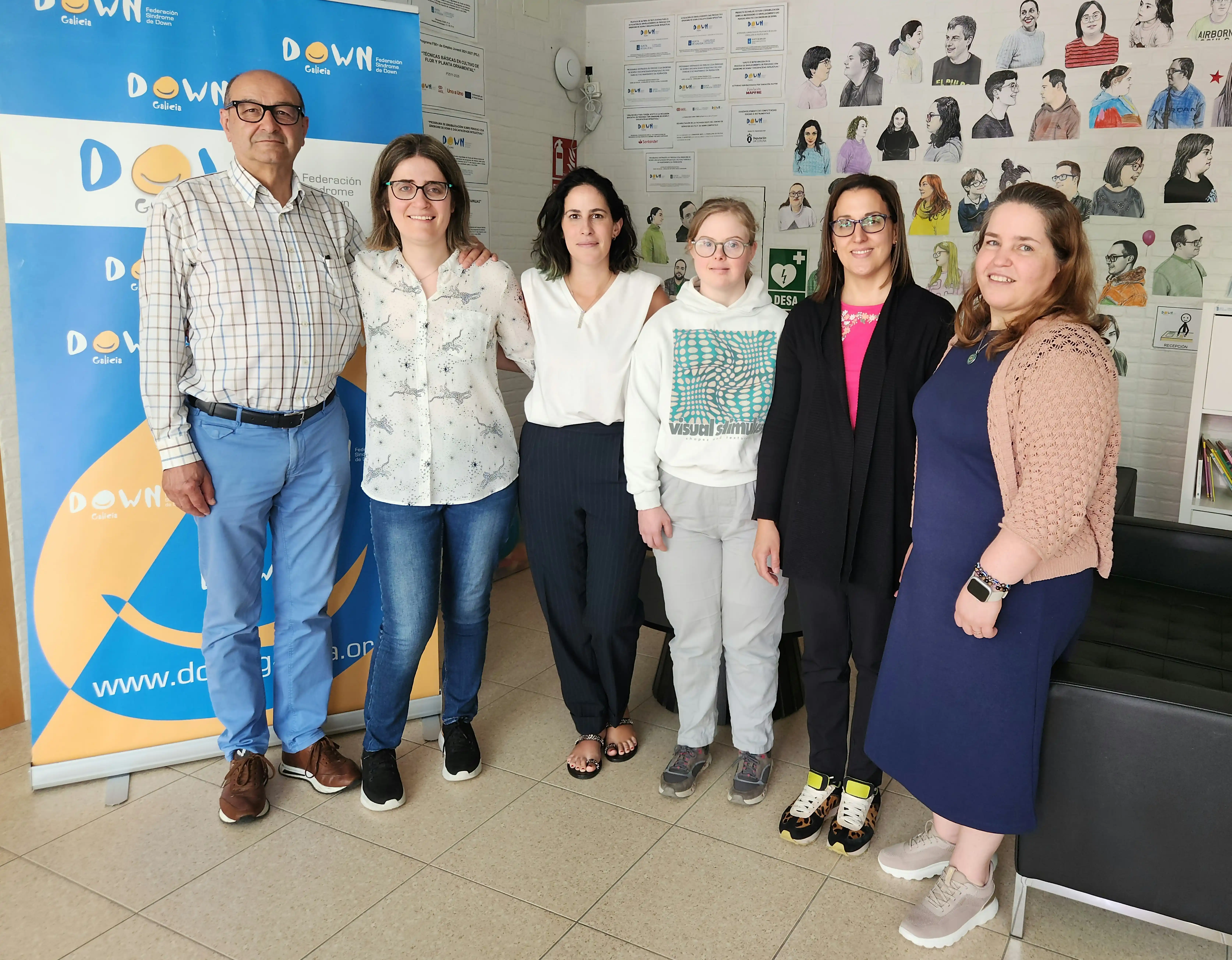 Group of six people posing in front of a banner of the Down Syndrome Federation of Galicia, indoors with walls decorated with illustrations of people with Down syndrome.
